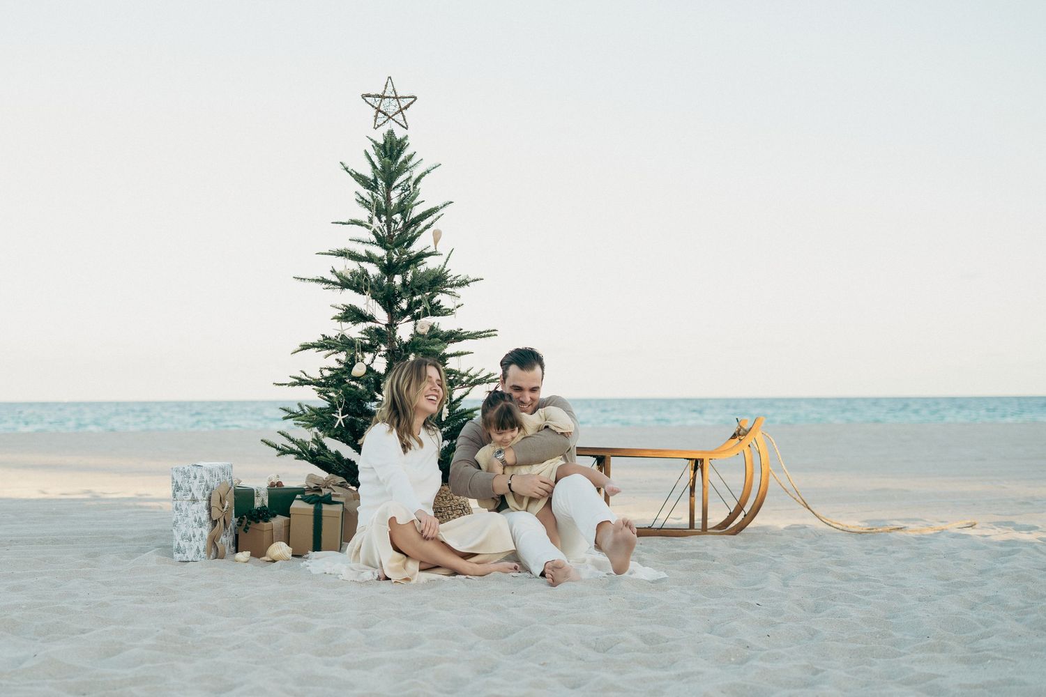 Couple sitting on beach sand by Christmas tree with gifts and wooden sled, celebrating holidays outdoors.