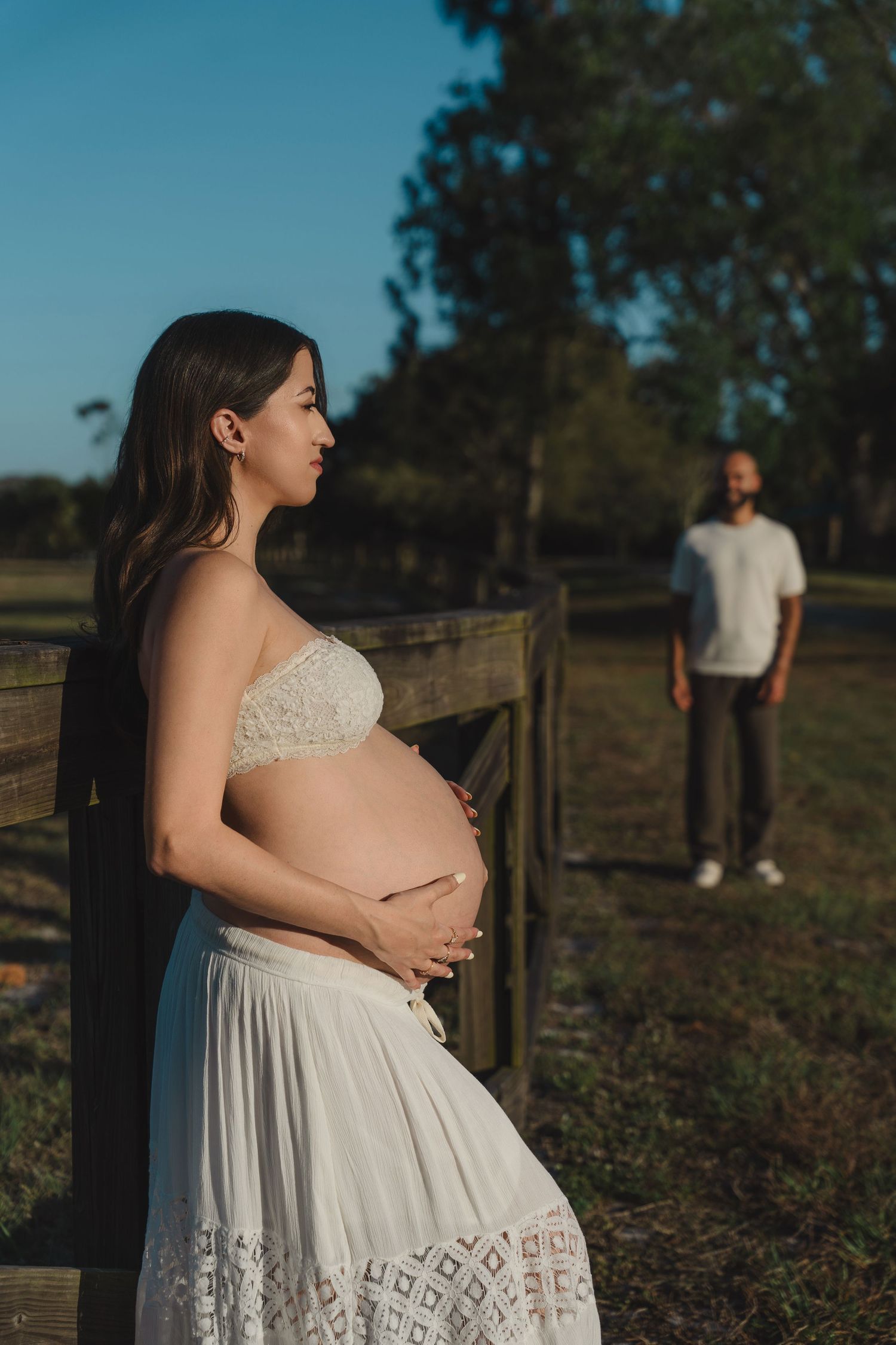 A woman in a flowing white lace dress cradles her pregnant belly in a rustic outdoor setting during golden hour.