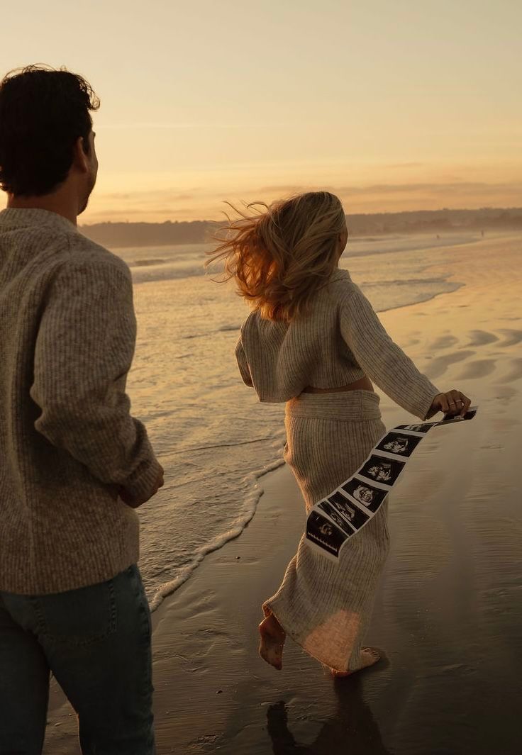 Couple walking on beach at sunset, woman holding ultrasound photos, maternity announcement by the ocean.