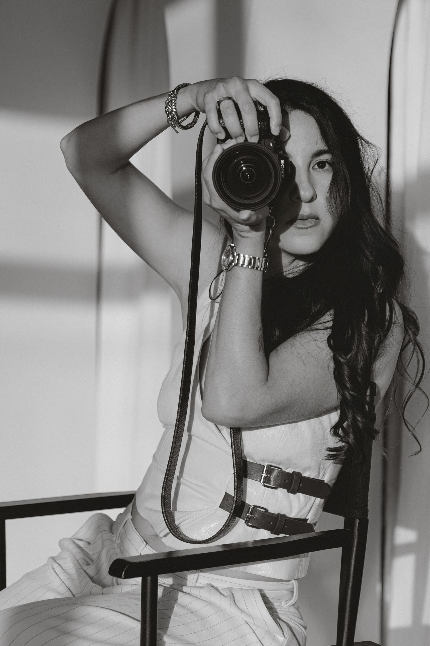 Black and white photo of a young woman with long hair holding a DSLR camera up to shoot, seated in a chair.
