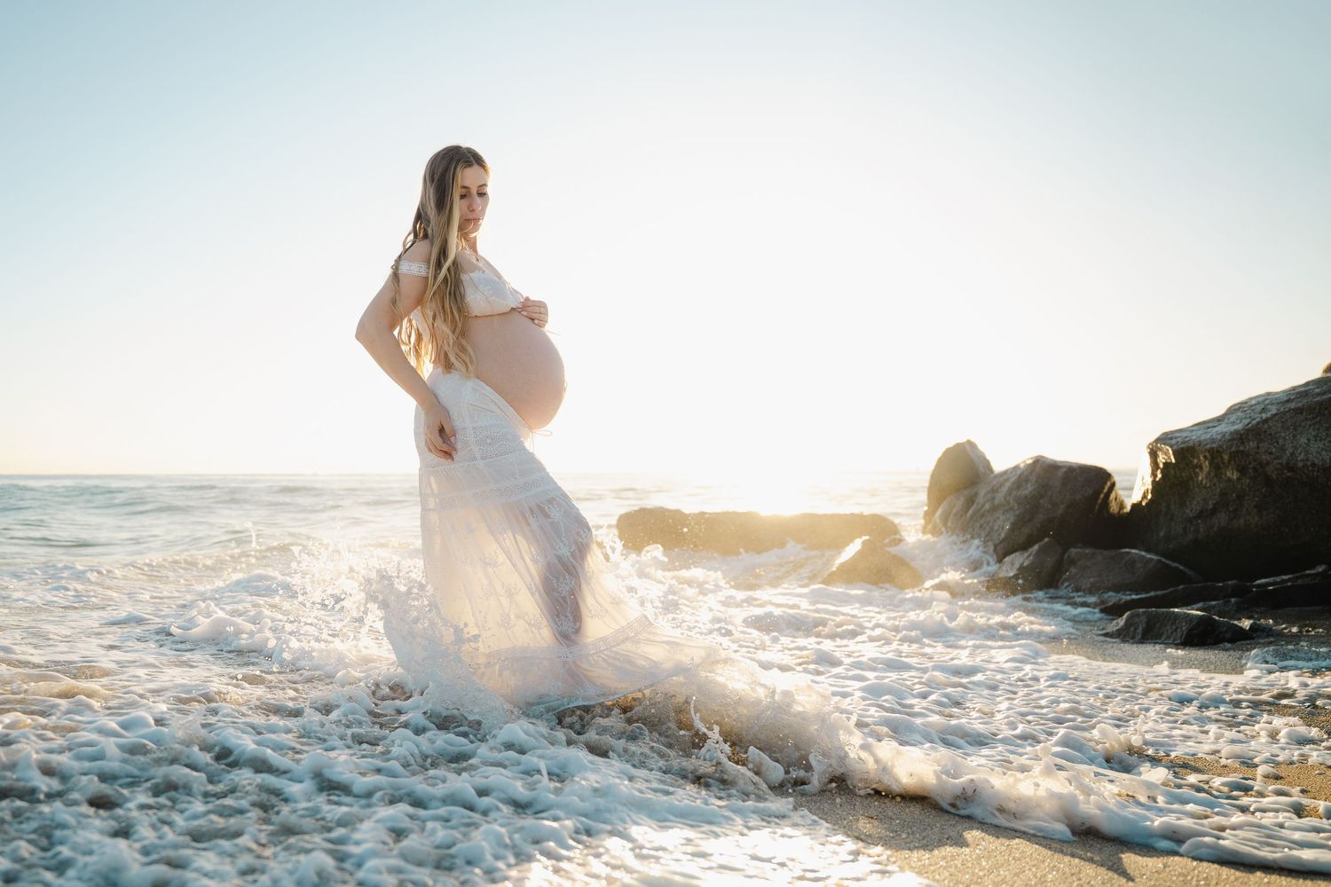A person in a flowing white dress dances through ocean waves at sunset creating ethereal splashing movements.