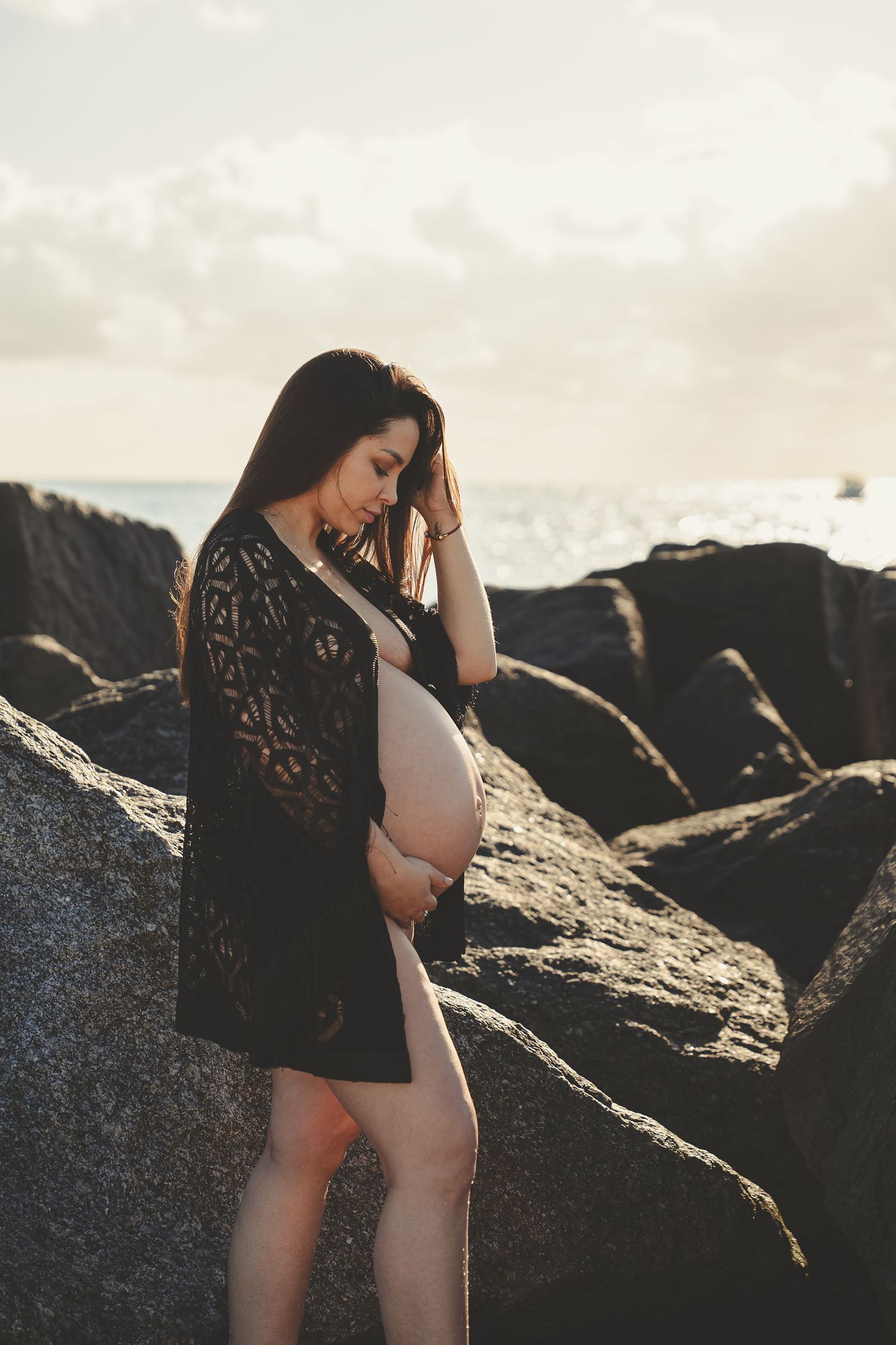 Artistic maternity photoshoot on dark rocky beach at sunset with lace dress details.