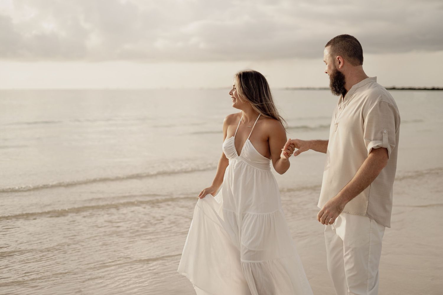 Couple in white outfits walking along a serene beach at sunset.