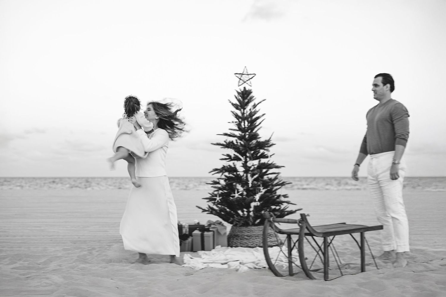 Black and white photo of a family celebrating Christmas on a beach with a decorated tree, gifts, and a sled.