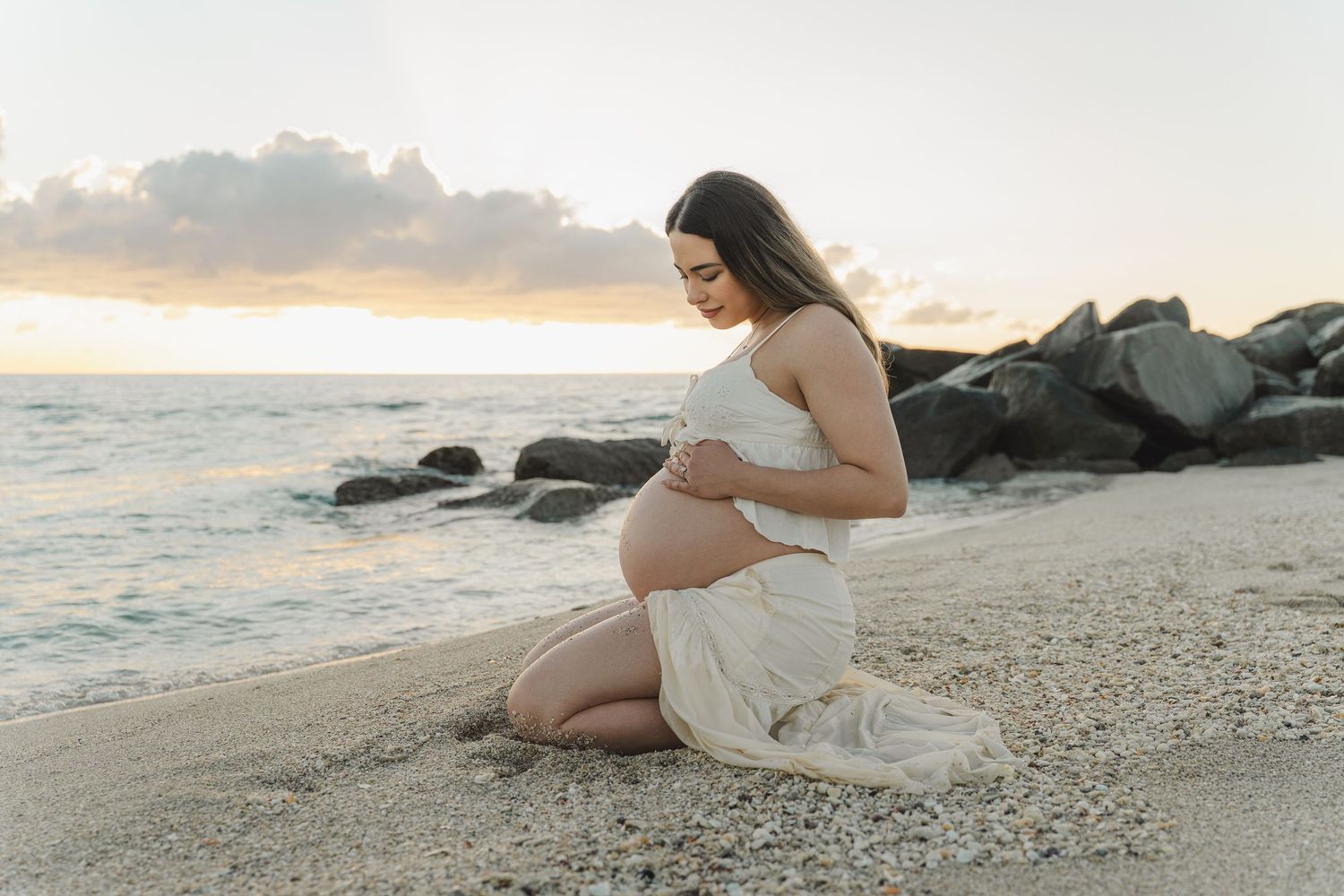 A person in a flowing white dress sits gracefully on a rocky beach at sunset, creating a serene coastal scene.