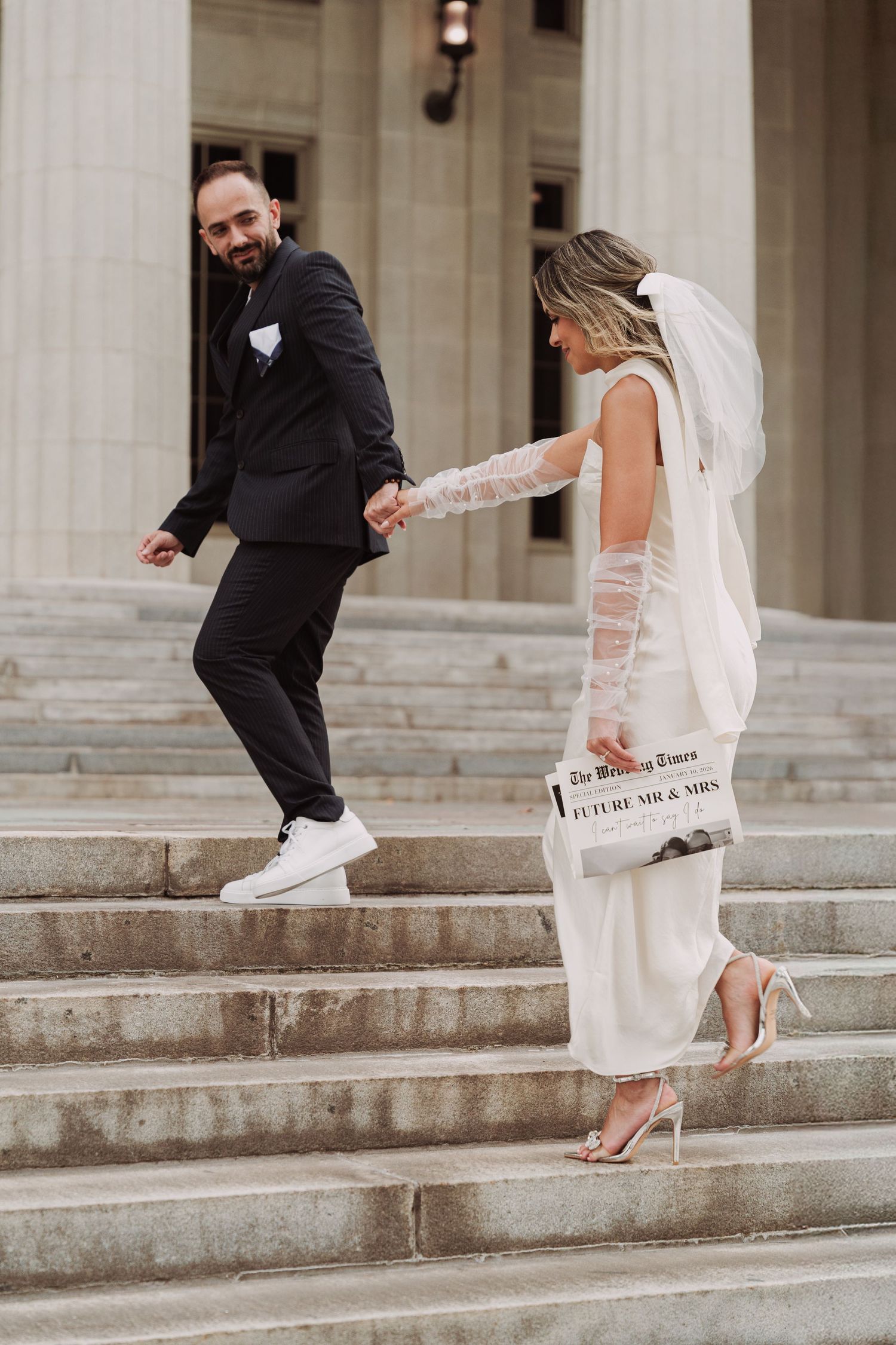 Wedding couple in formal attire playfully posing on grand stone steps of classical building.