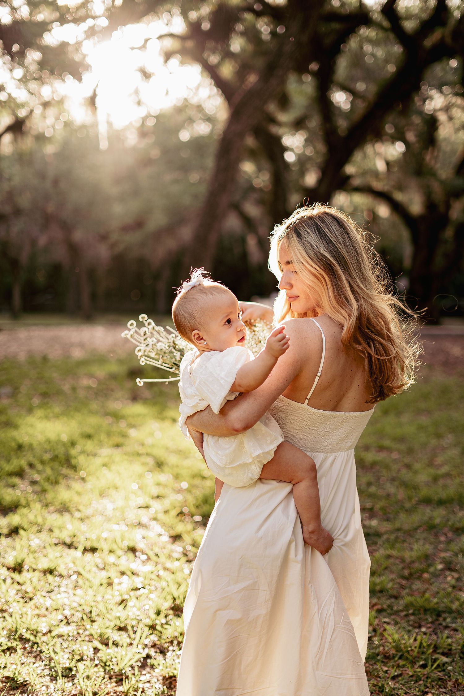 Mother and baby in white dresses playing in golden sunlight outdoors.