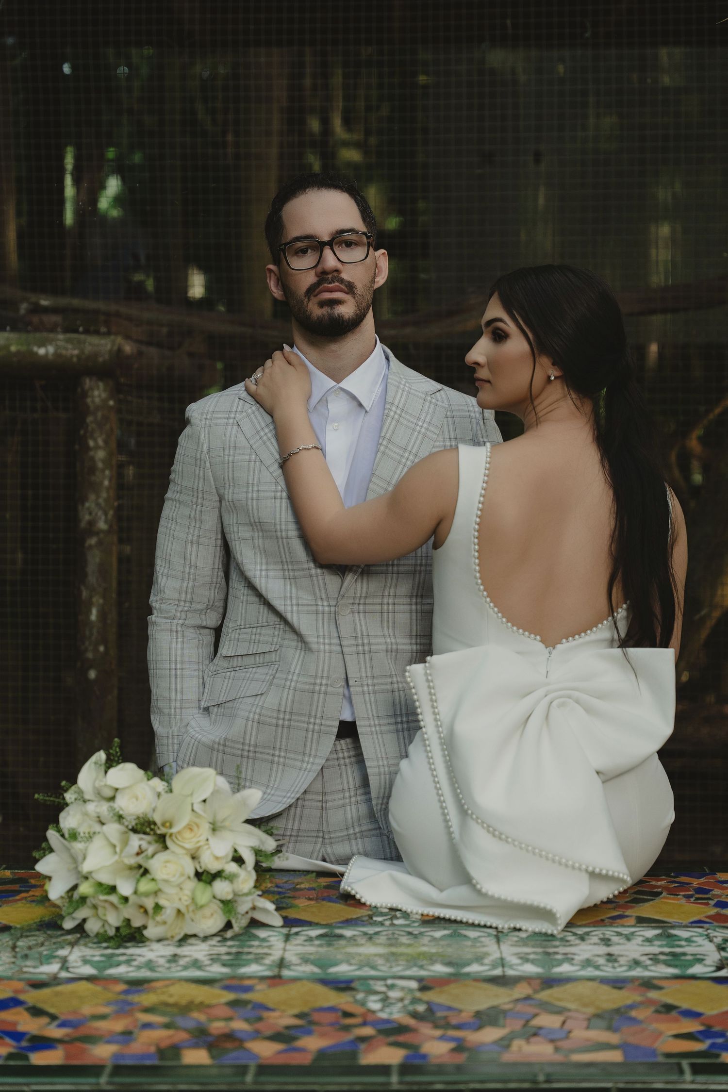 Bride in backless white gown with bow and groom in plaid suit sit together at mosaic table with white floral bouquet.
