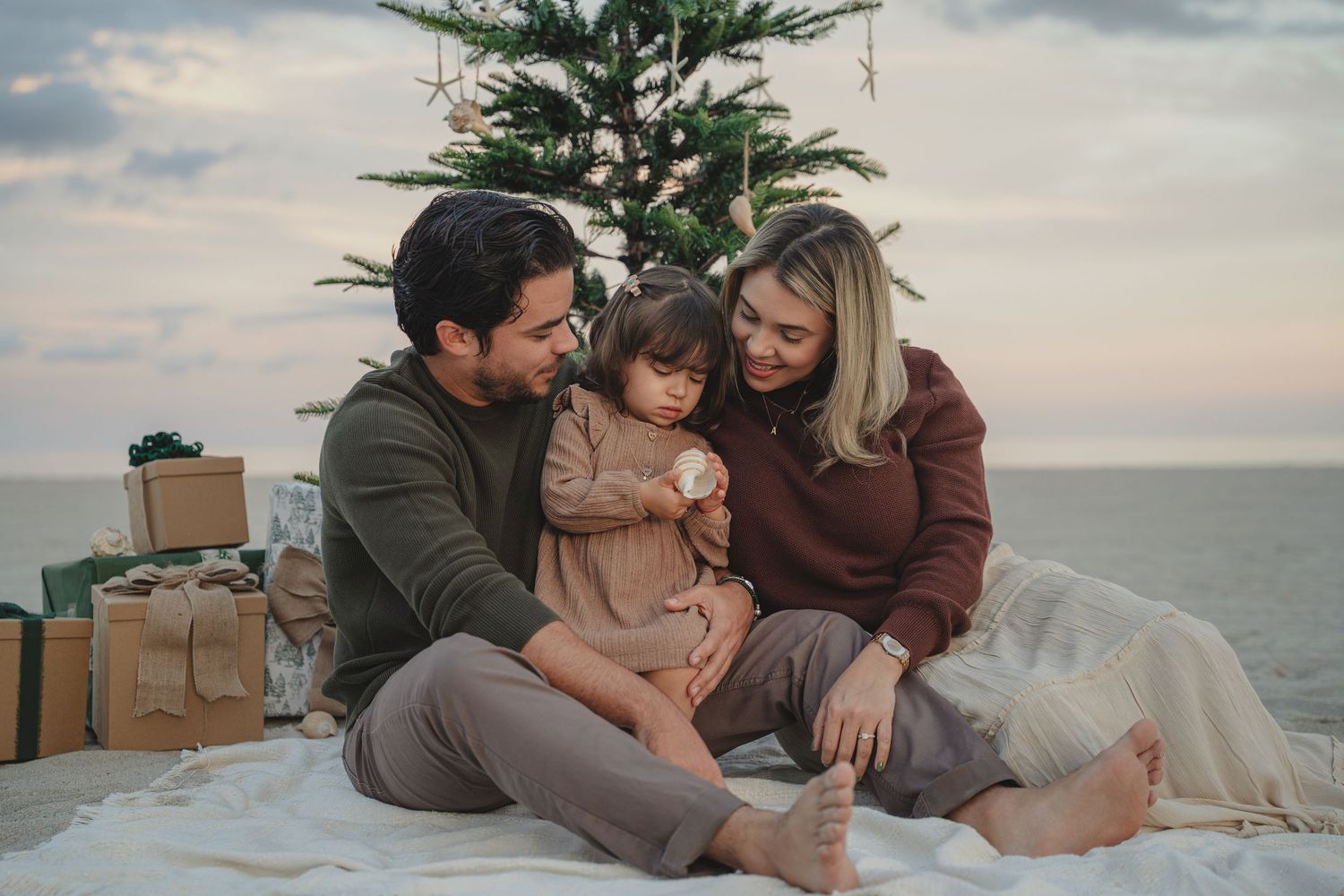 Family sitting on beach blanket by Christmas tree with gifts, celebrating holiday season outdoors near ocean.