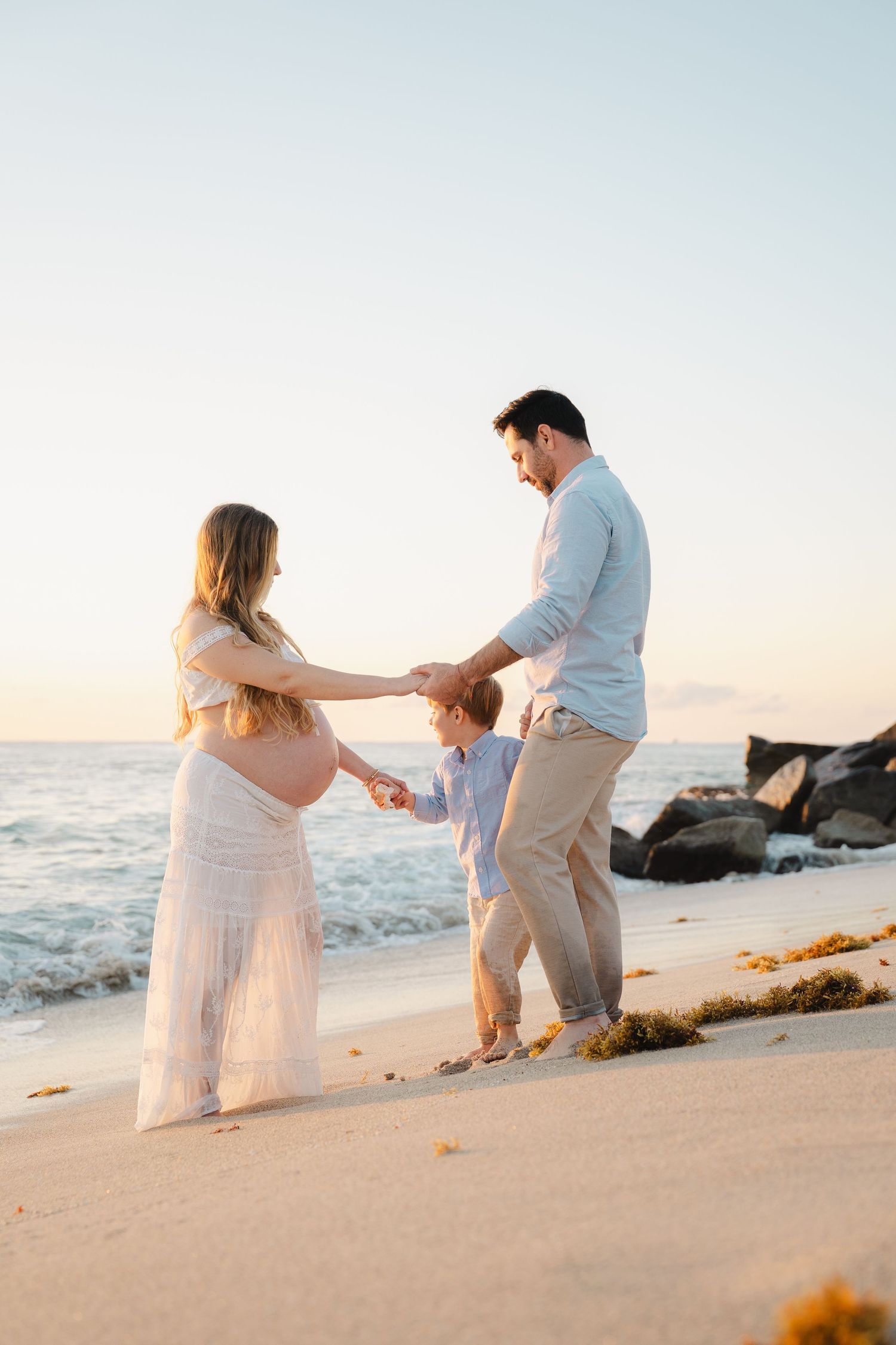 Pregnant woman in white dress holds hands with young son and husband on sandy beach at sunset.