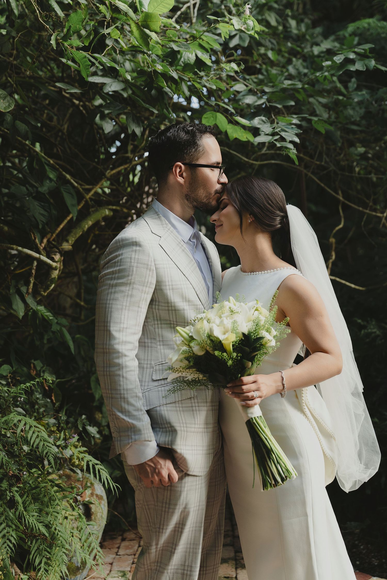A groom in a plaid suit kisses his bride wearing a white gown and veil, holding a bouquet in a lush garden.