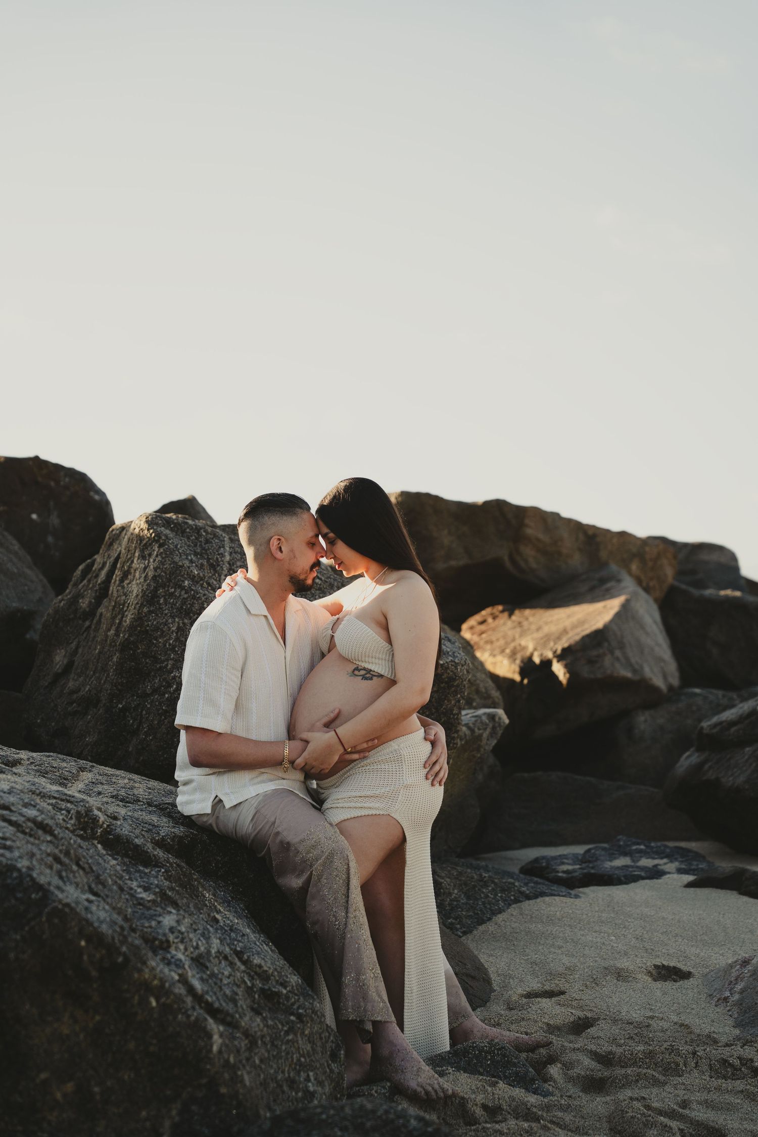 Romantic maternity photos on rocky beach during golden hour.