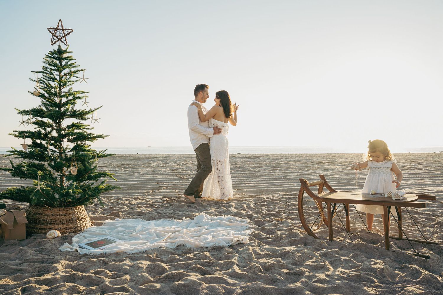 Couple embracing on sandy beach with Christmas tree, sled, and child nearby during golden hour.