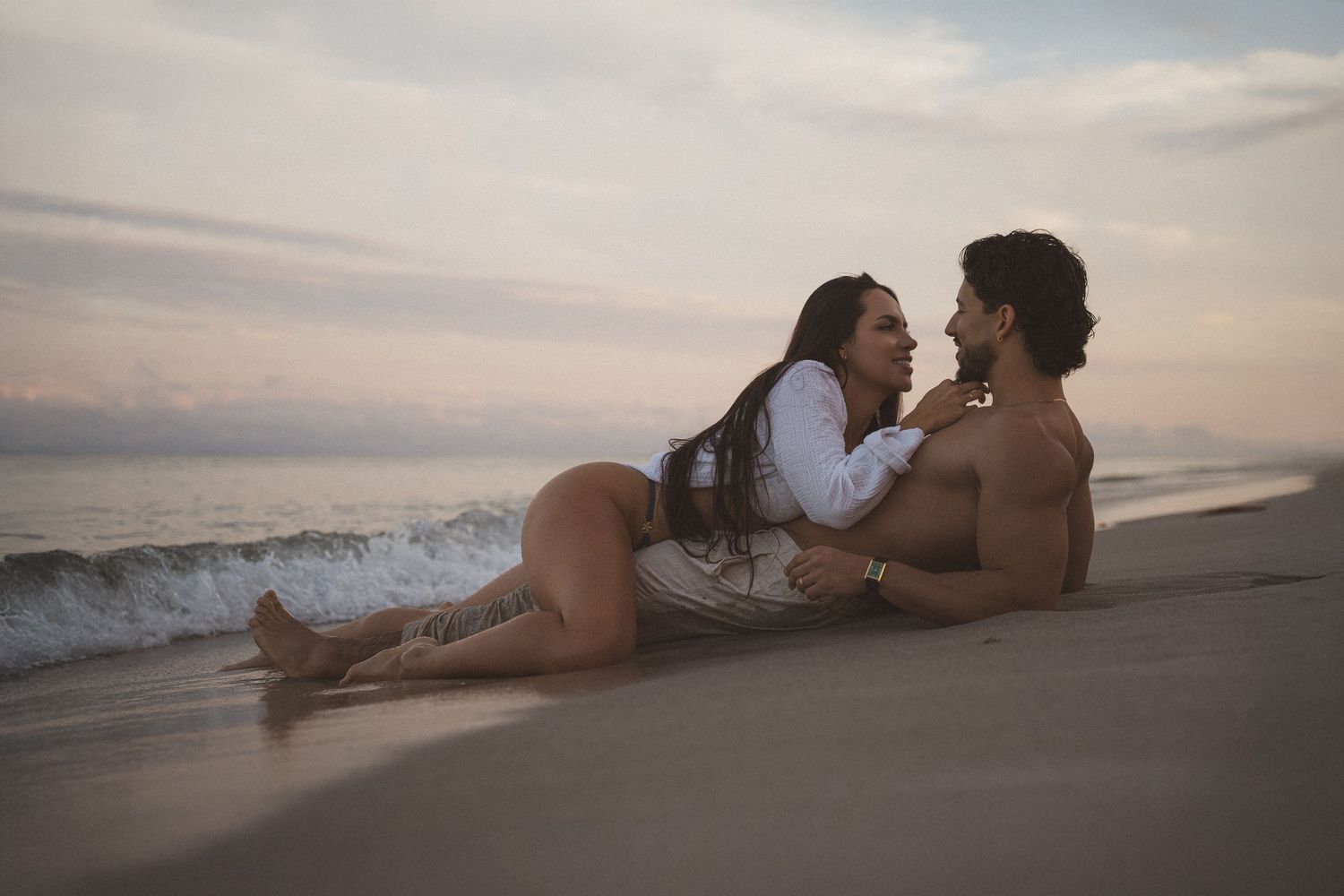 Romantic couple sitting together on a sandy beach at sunset with ocean waves in the background.