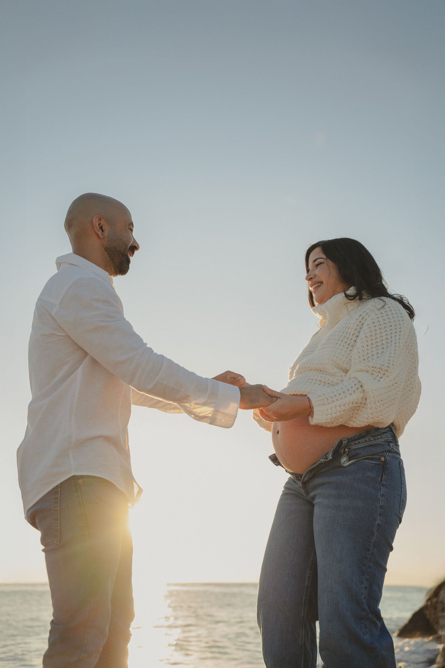 Couple holding hands at sunset by the ocean, pregnant woman in white sweater and jeans smiling at her partner.