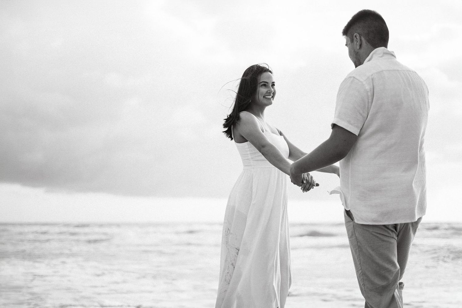 Black and white beach engagement photos with couple in white outfits against ocean backdrop.