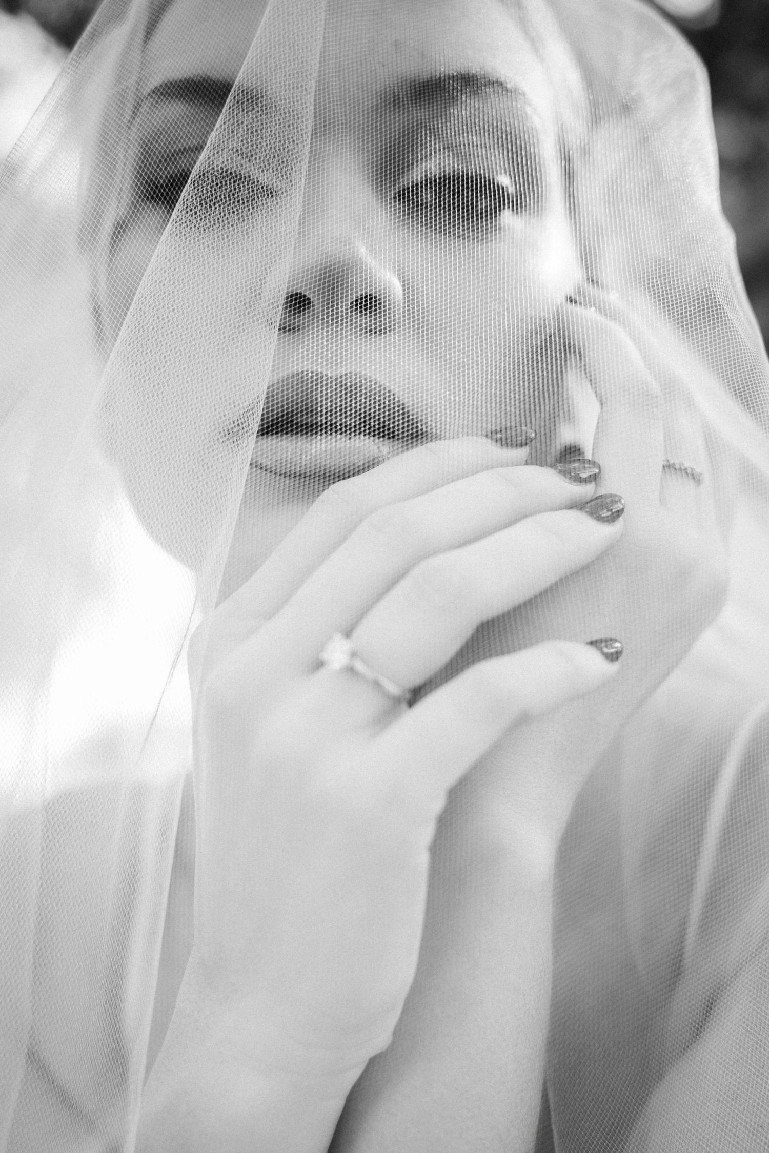 Black and white portrait of a bride wearing a sheer veil, holding her hand to her face, wearing a diamond ring.
