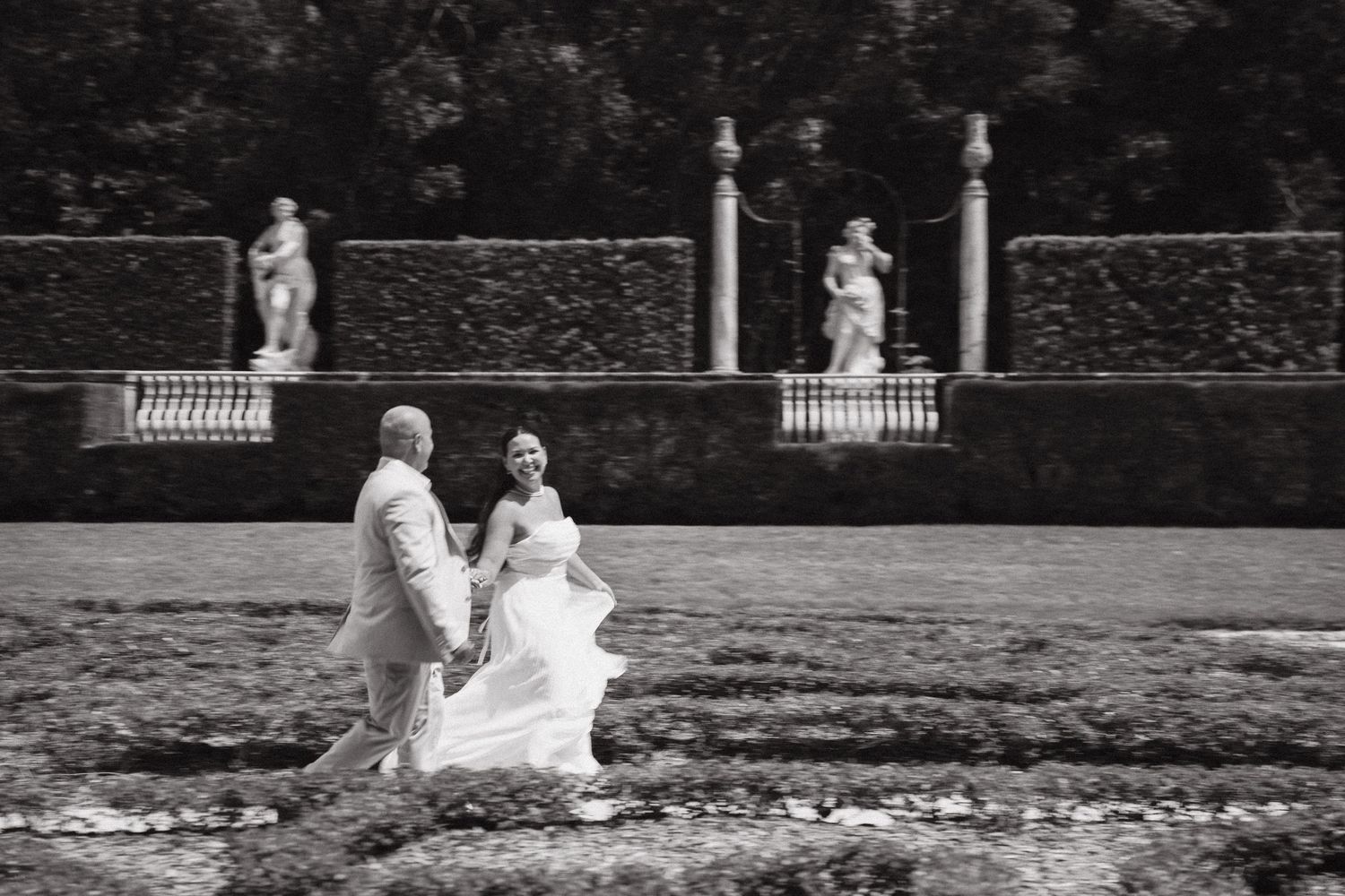 Black and white photo of a bride and groom walking hand in hand in an elegant garden with classical statues.