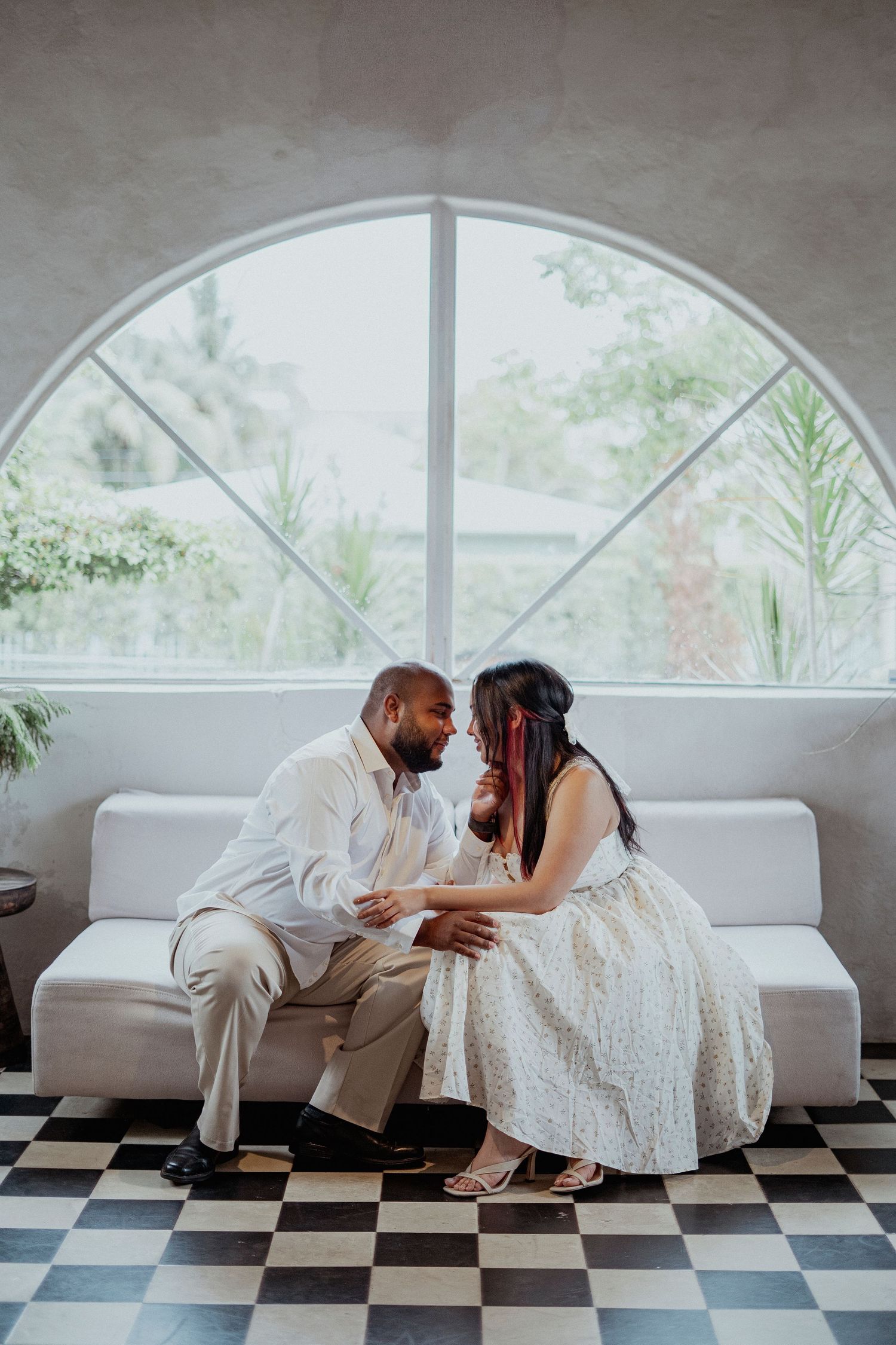 Couple in white attire posing on checkered floor near arched windows.