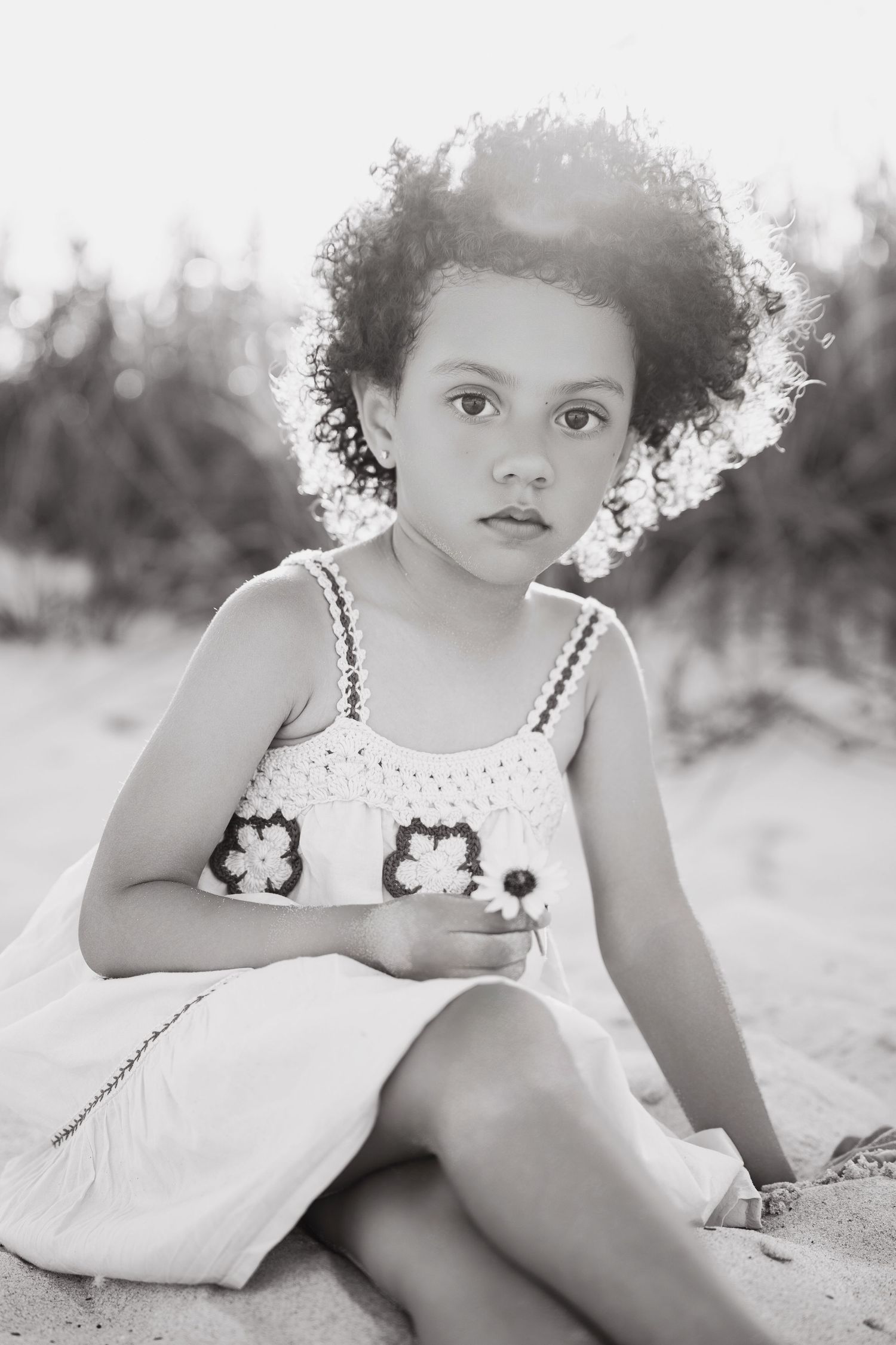 Black and white portrait of a young girl with curly hair sitting on sand, wearing a white dress and holding a flower.