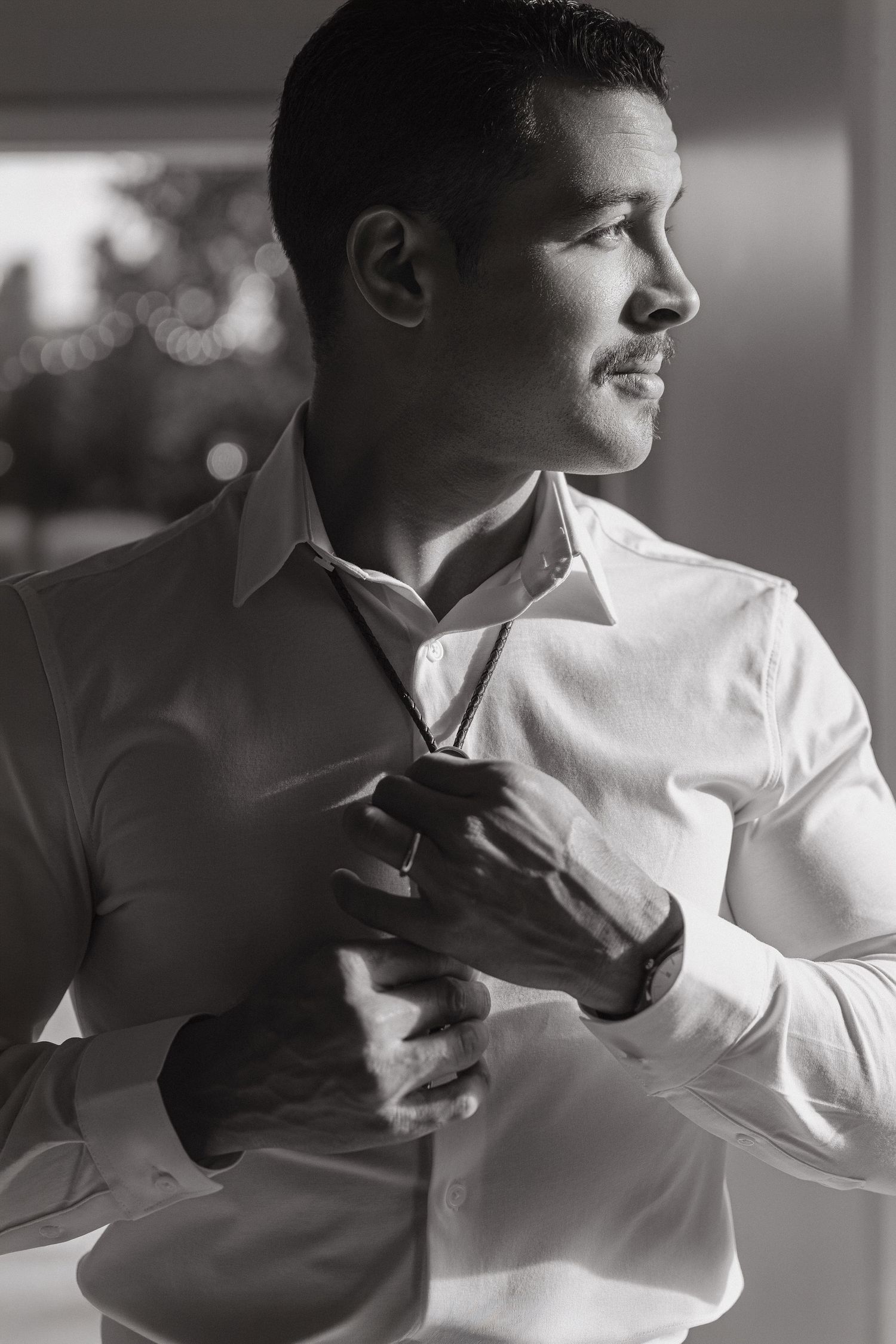 Black and white photo of a man in a white dress shirt adjusting his tie, looking away thoughtfully.