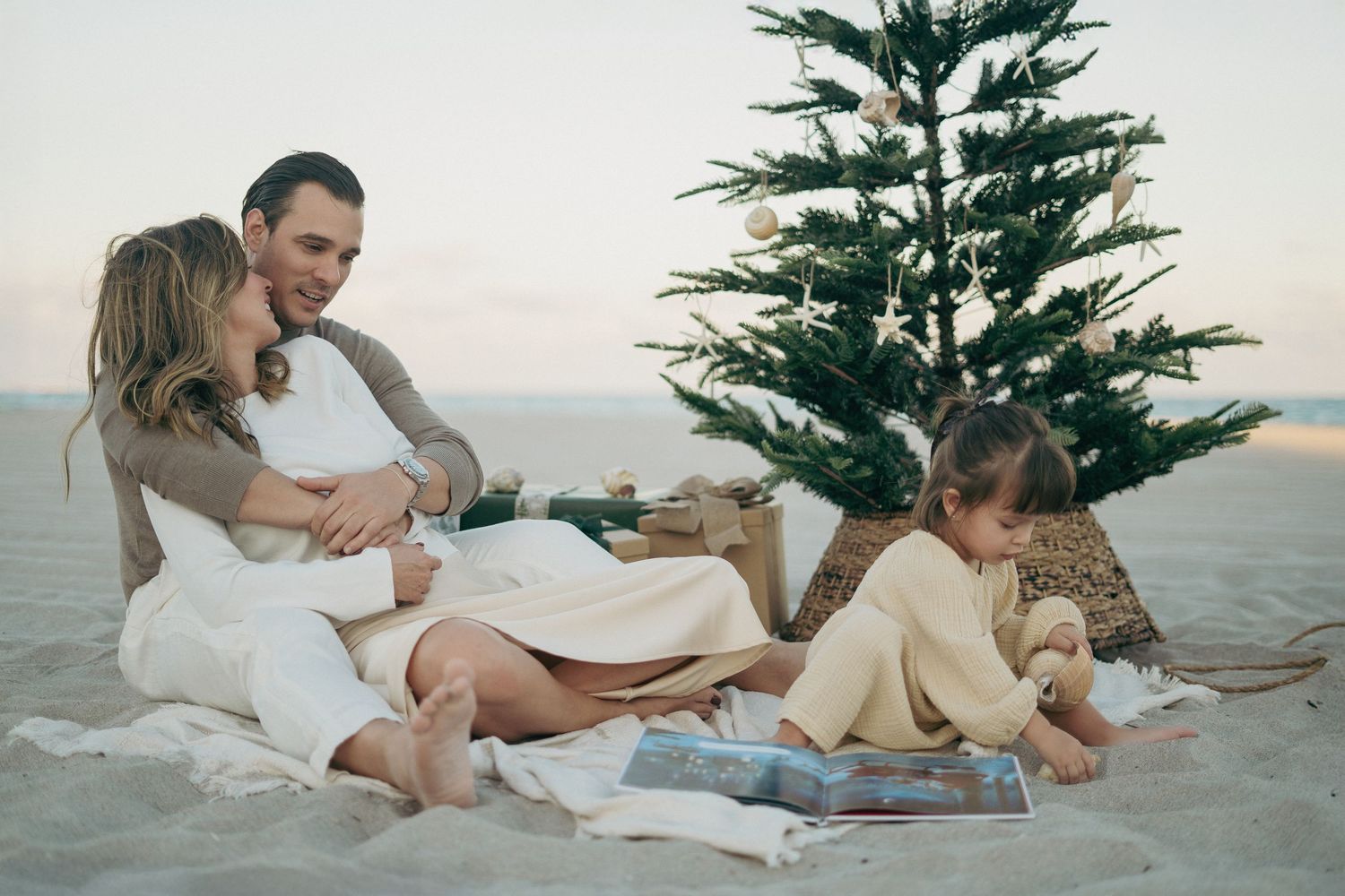 Family celebrating Christmas on a sandy beach beside a decorated tree with wrapped gifts at sunset.