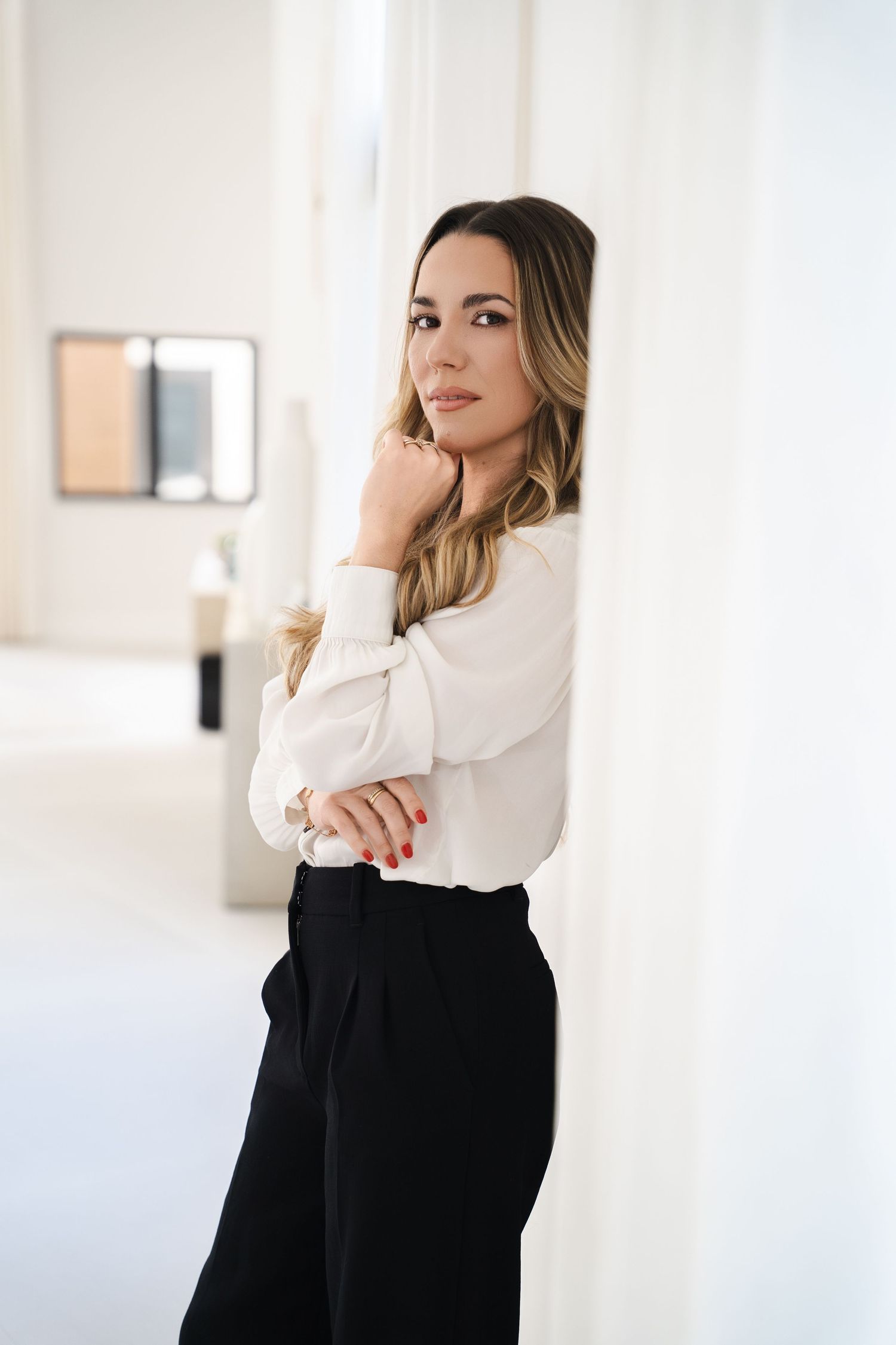 Woman in white blouse and black trousers leaning against a white wall in a bright modern interior.