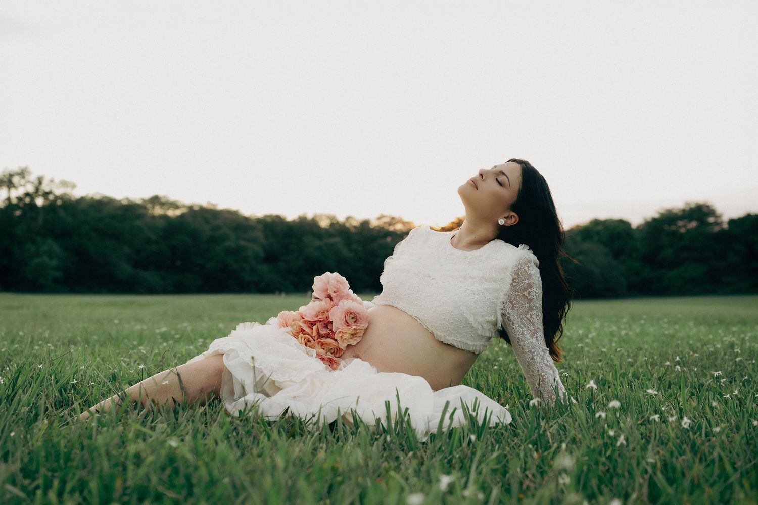 Pregnant woman in white lace dress reclines on green grass holding pink flowers during golden hour maternity photoshoot.