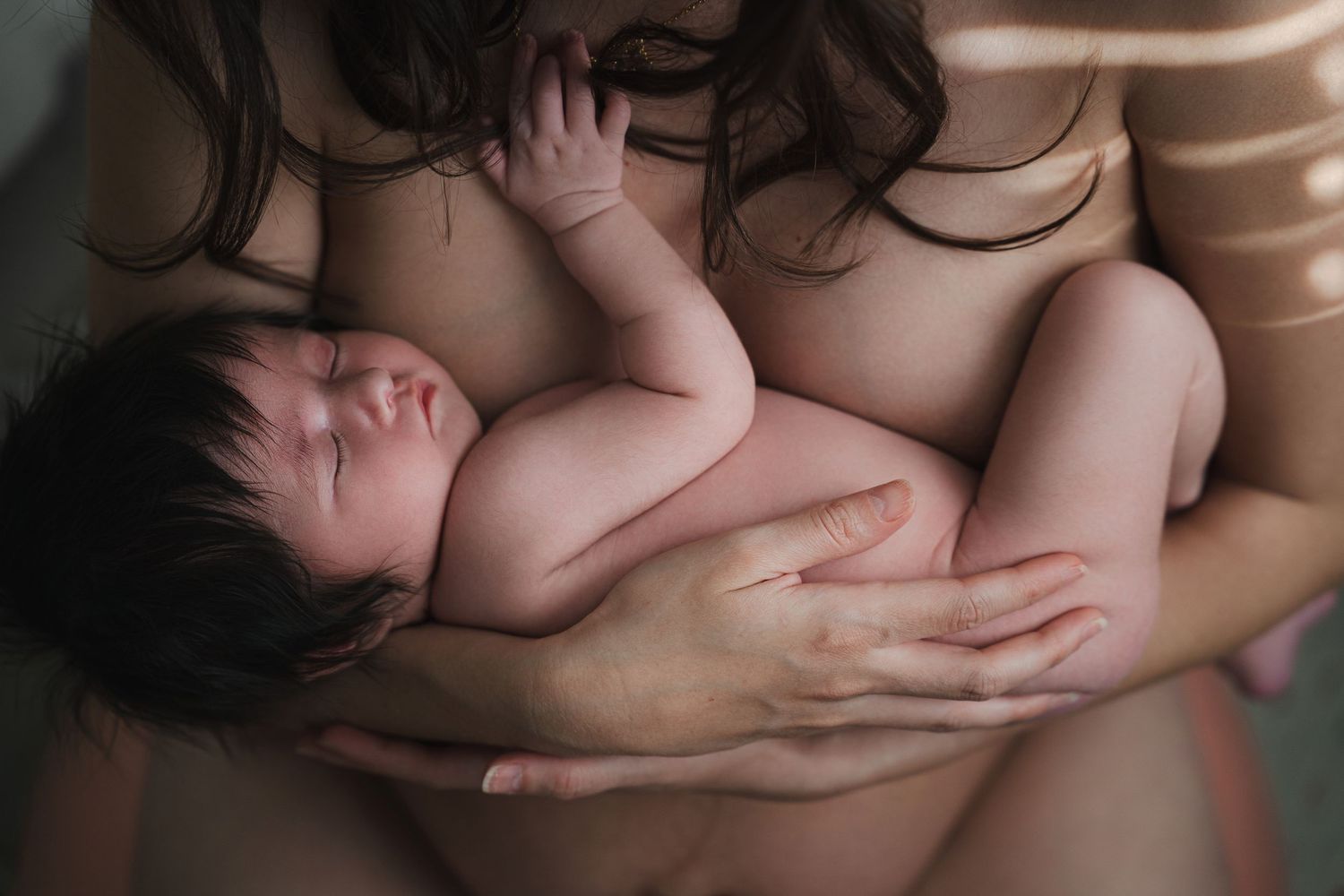 A mother tenderly cradles her sleeping newborn baby against her chest in a soft, intimate portrait.