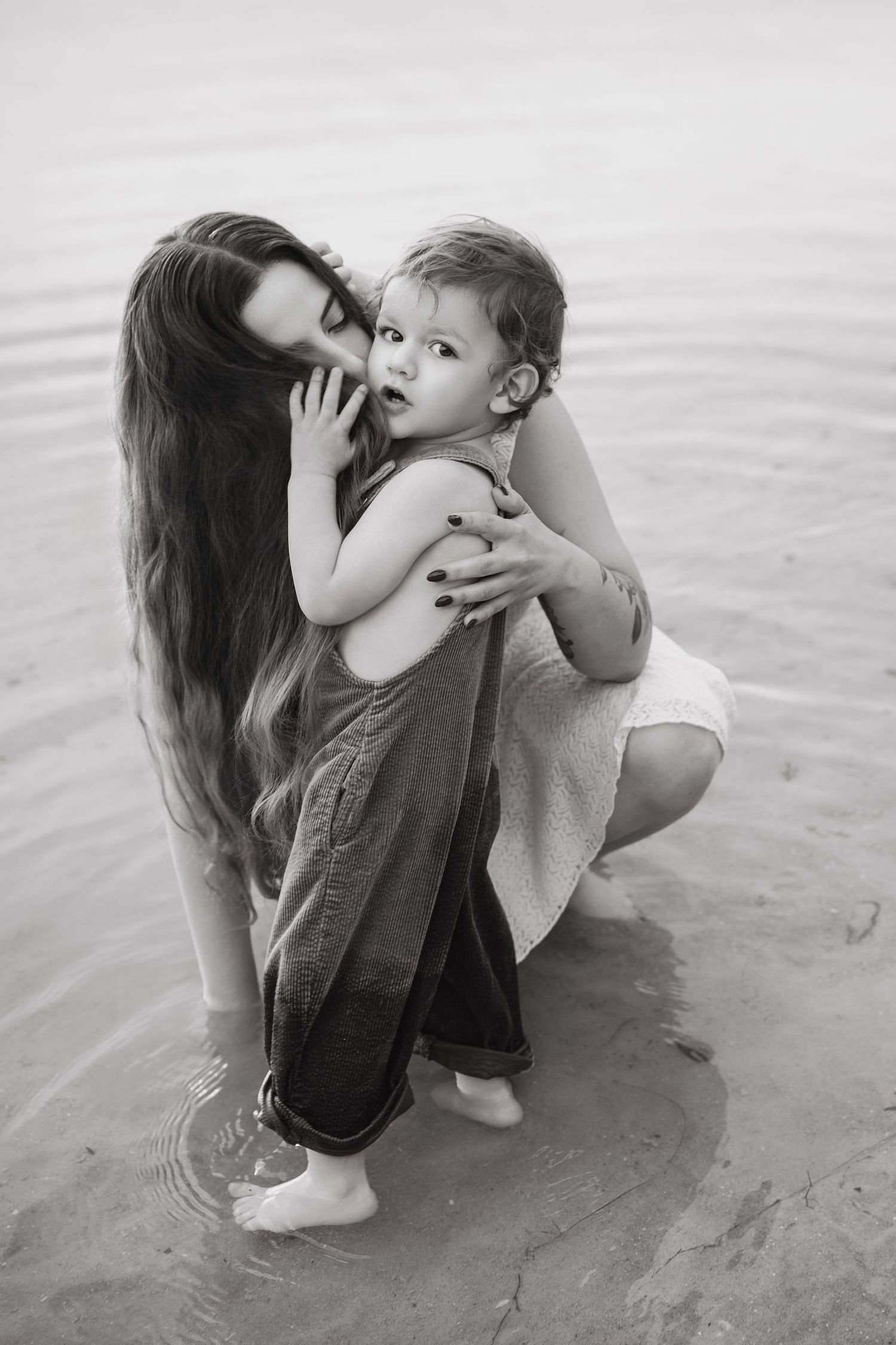 Black and white photo of a mother hugging her toddler child while crouching in shallow water near a beach.