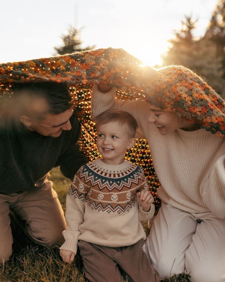 Family of three sharing a warm moment under a blanket at golden hour sunset outdoors.