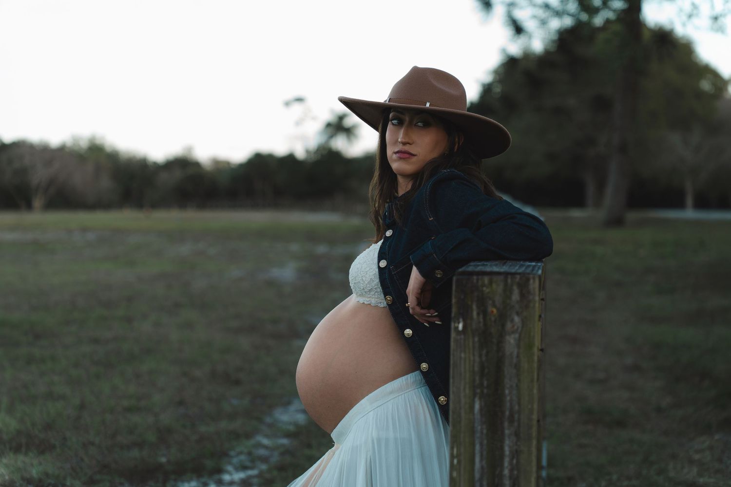 A pregnant woman in a flowing white dress stands in a sunlit field during golden hour.