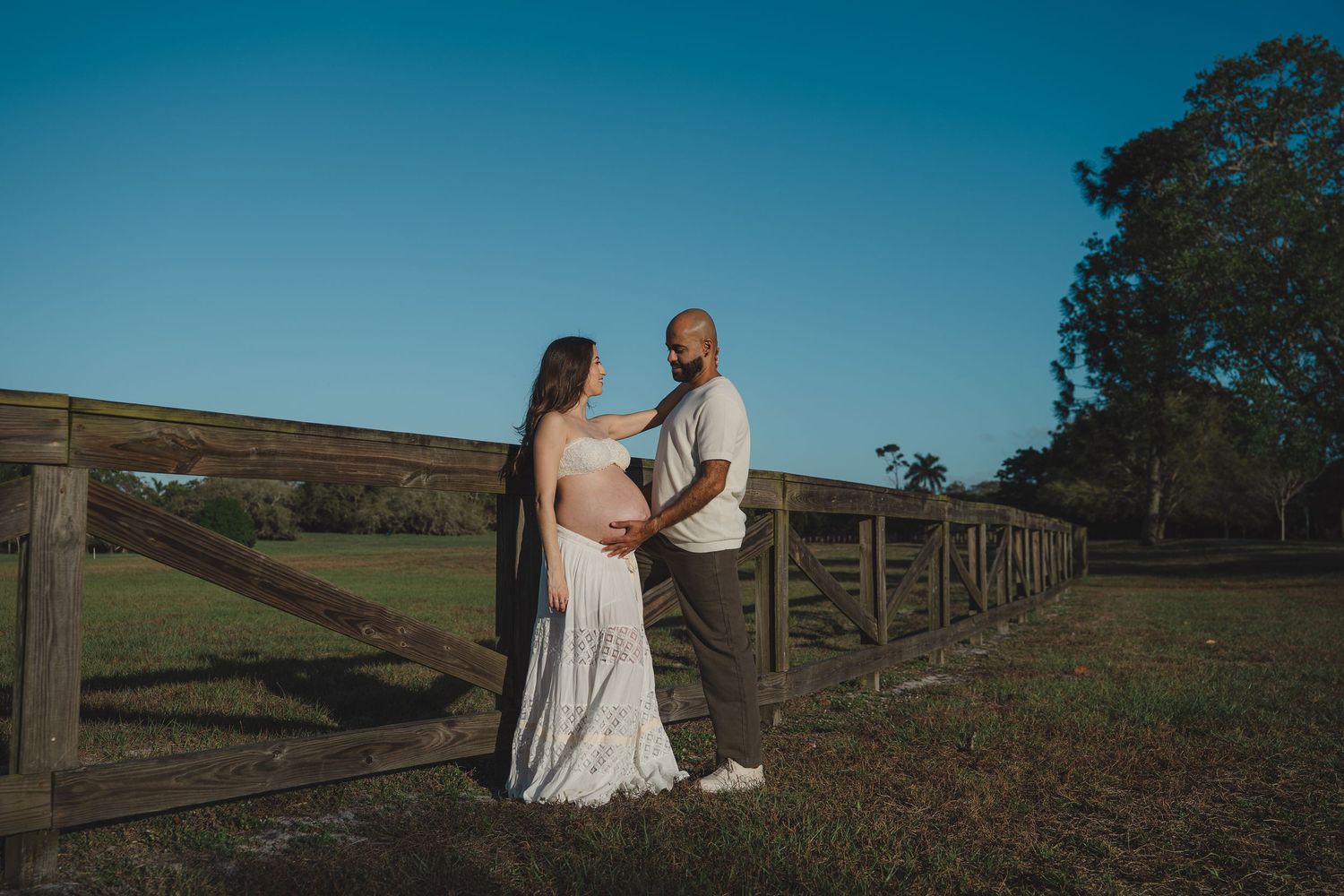 A couple shares an intimate moment along a rustic wooden fence at sunset on a countryside hillside.