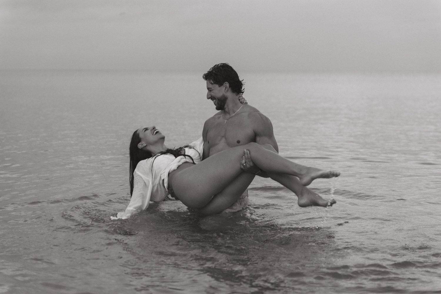 Black and white photo of a couple embracing romantically in shallow ocean water on a cloudy day.