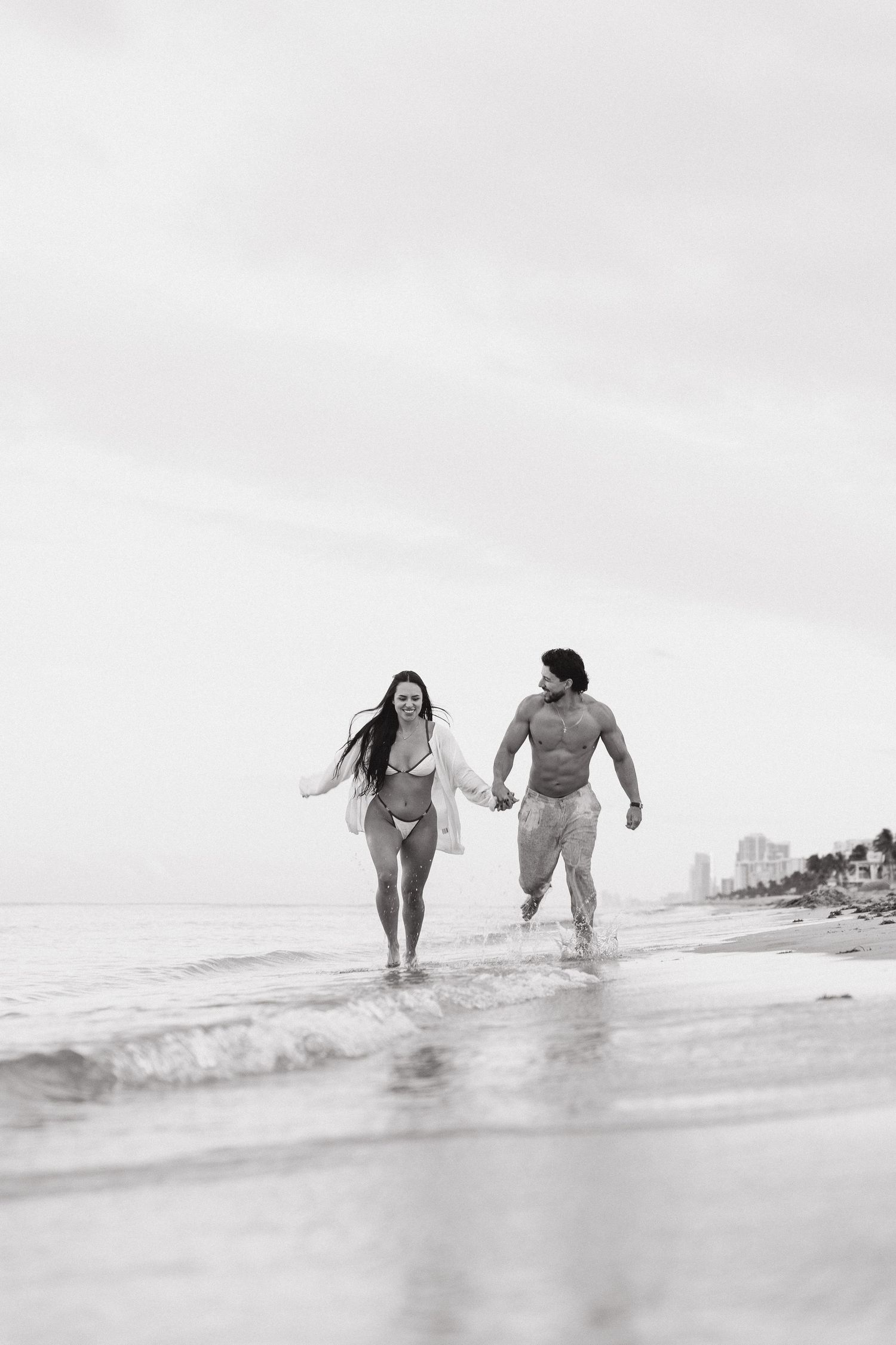 Black and white photo of a couple holding hands and running along the ocean shoreline in swimwear.