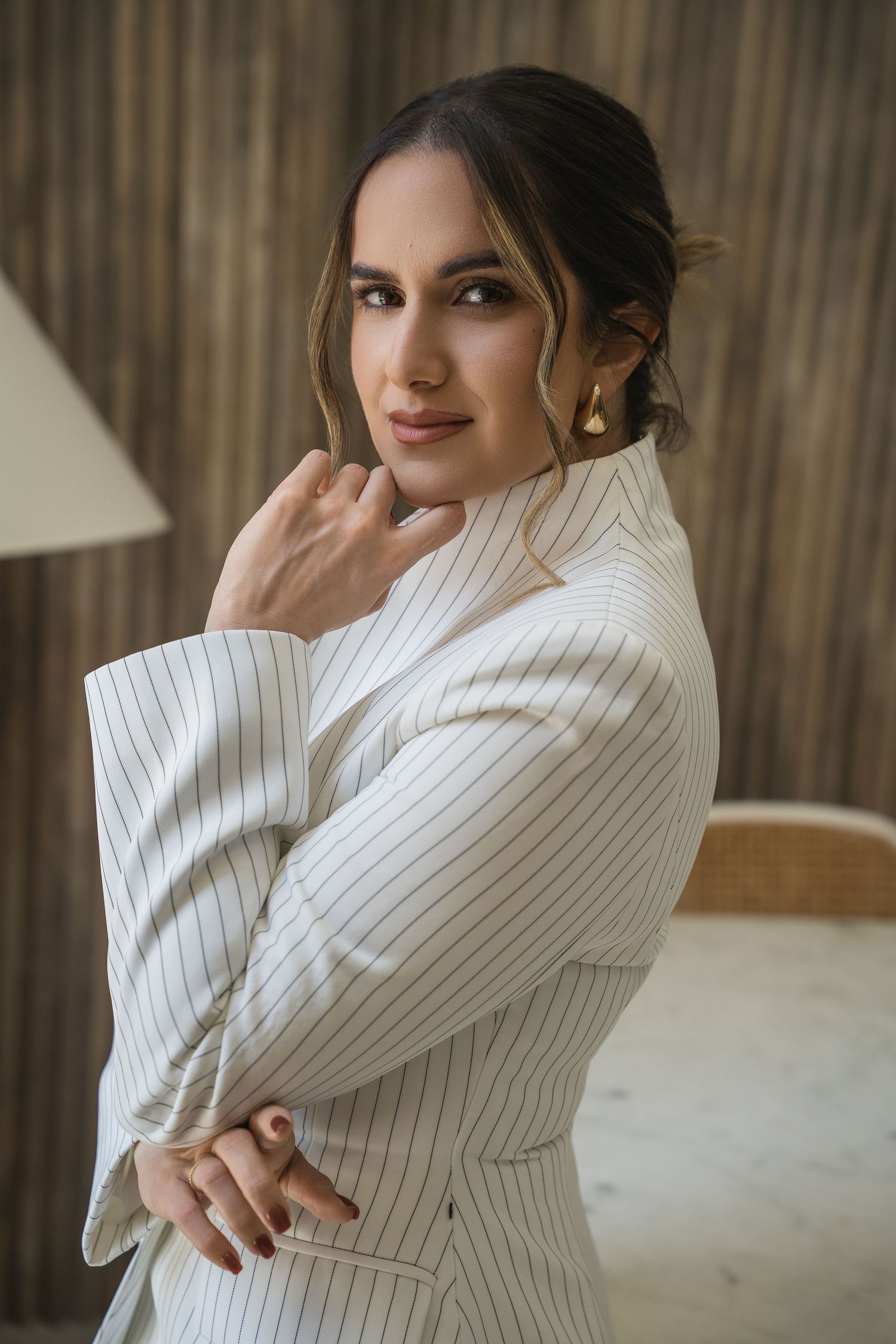 Woman in white pinstripe blazer poses elegantly with arms crossed, wearing gold earrings against a wooden backdrop.