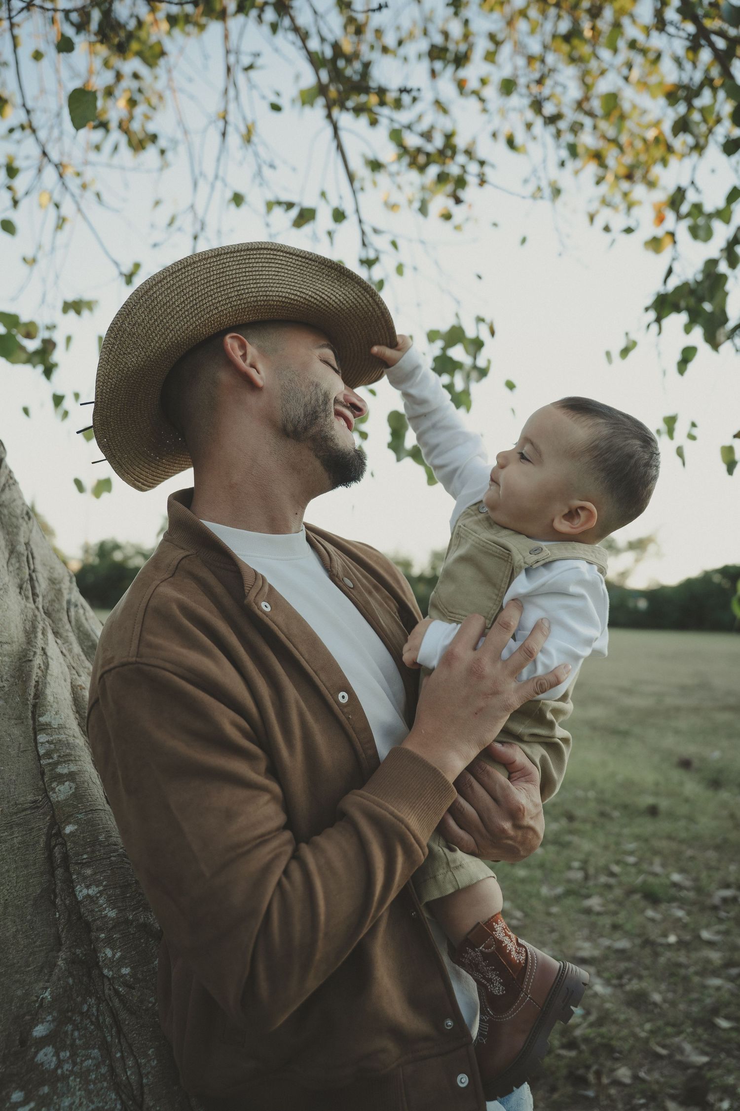 Tender parent-child moment captured under tree branches in warm evening light.
