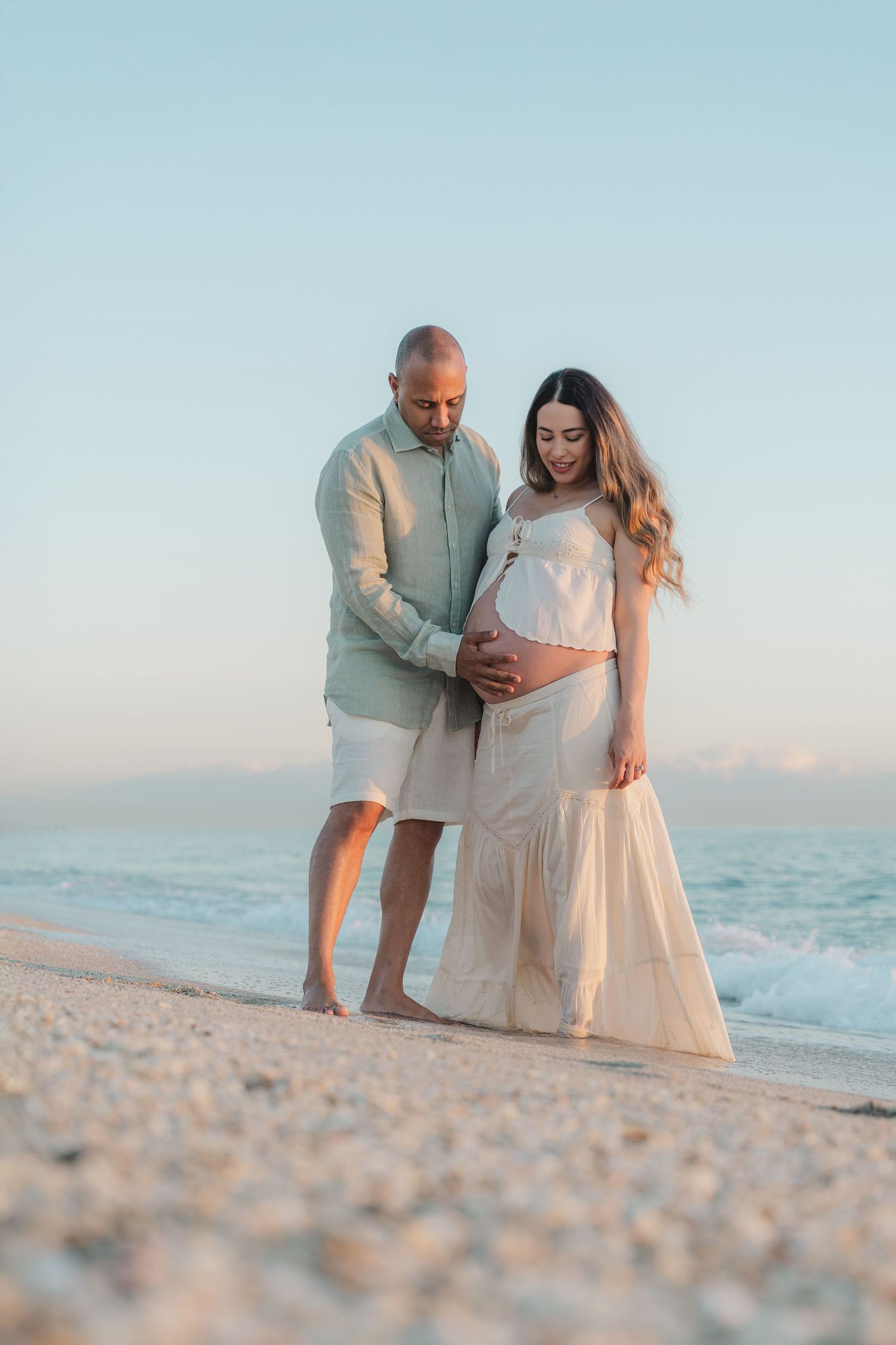 A couple tenderly cradles a pregnant belly on a serene beach at golden hour, wearing soft neutral tones.