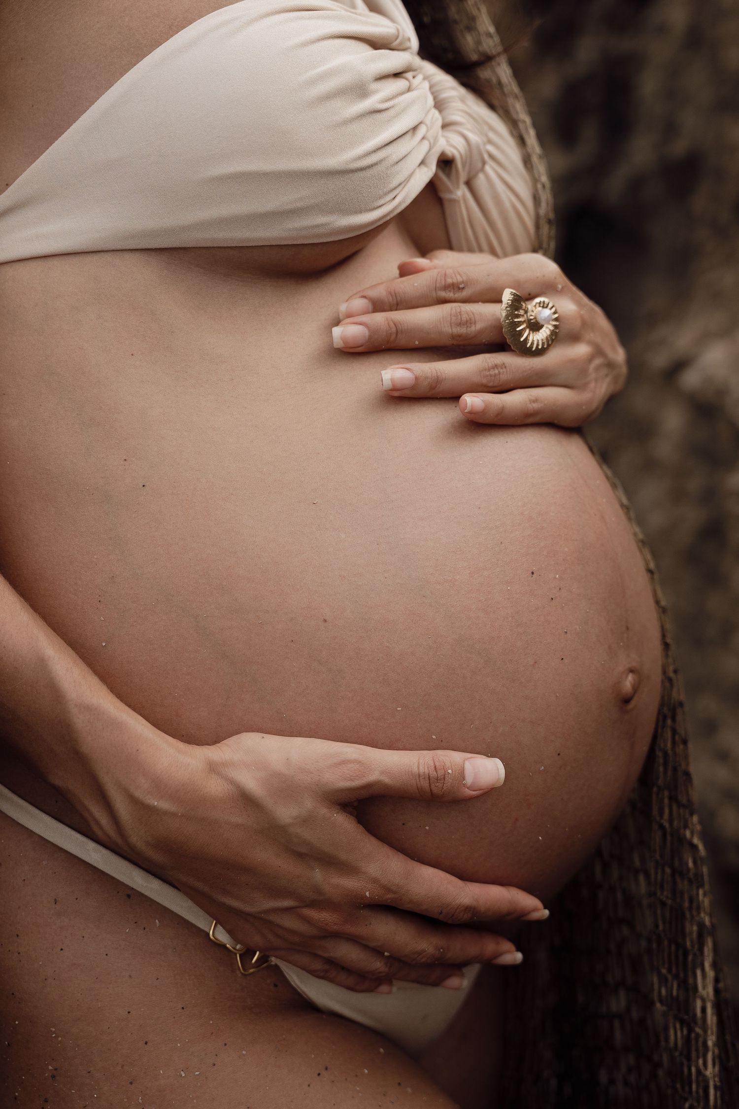 Pregnant woman in white bikini top cradling her baby bump with both hands, wearing a gold flower ring.