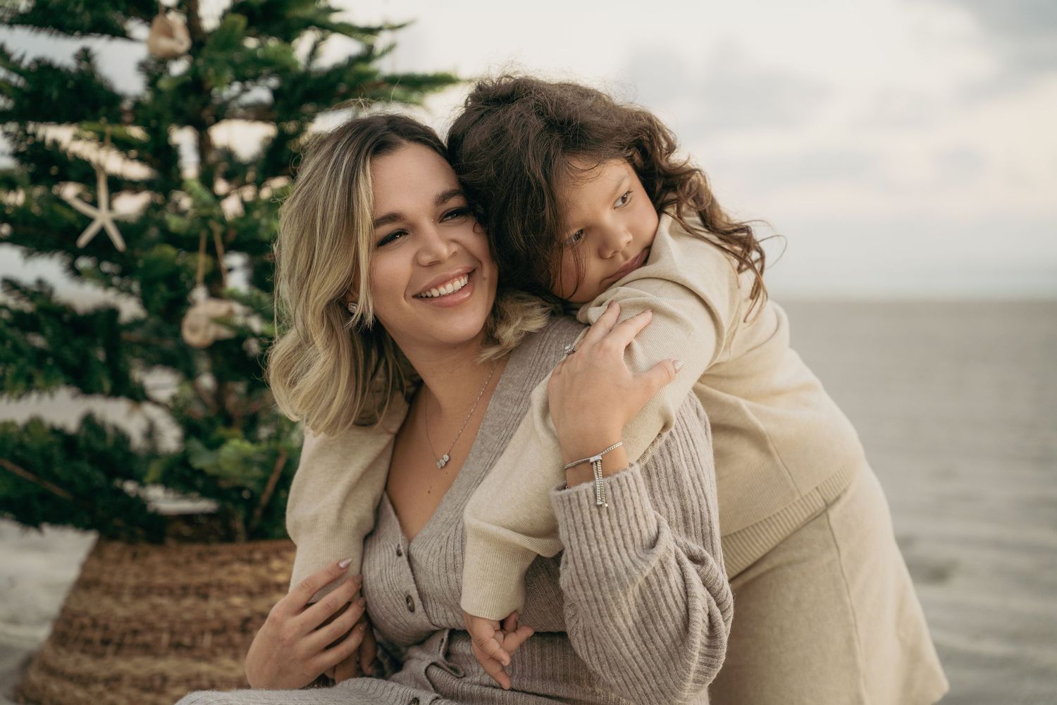 A smiling woman and young girl share a warm hug near a Christmas tree by the ocean.