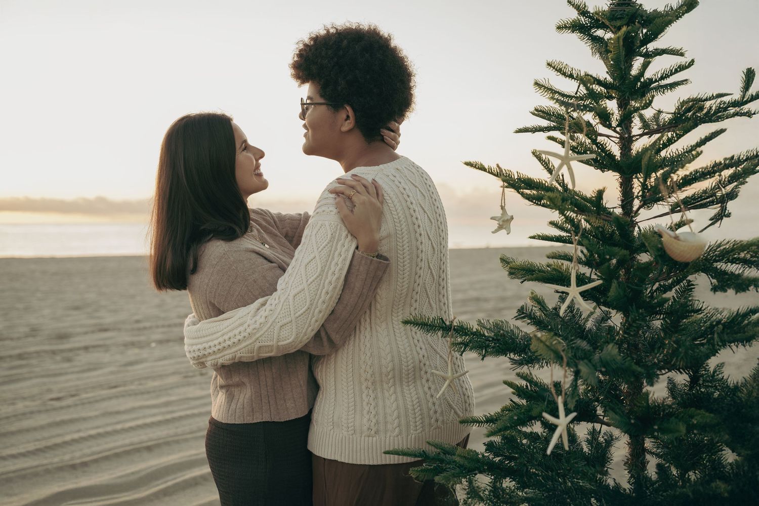 A couple embraces near a decorated Christmas tree on a beach at sunset, wearing cozy knit sweaters.