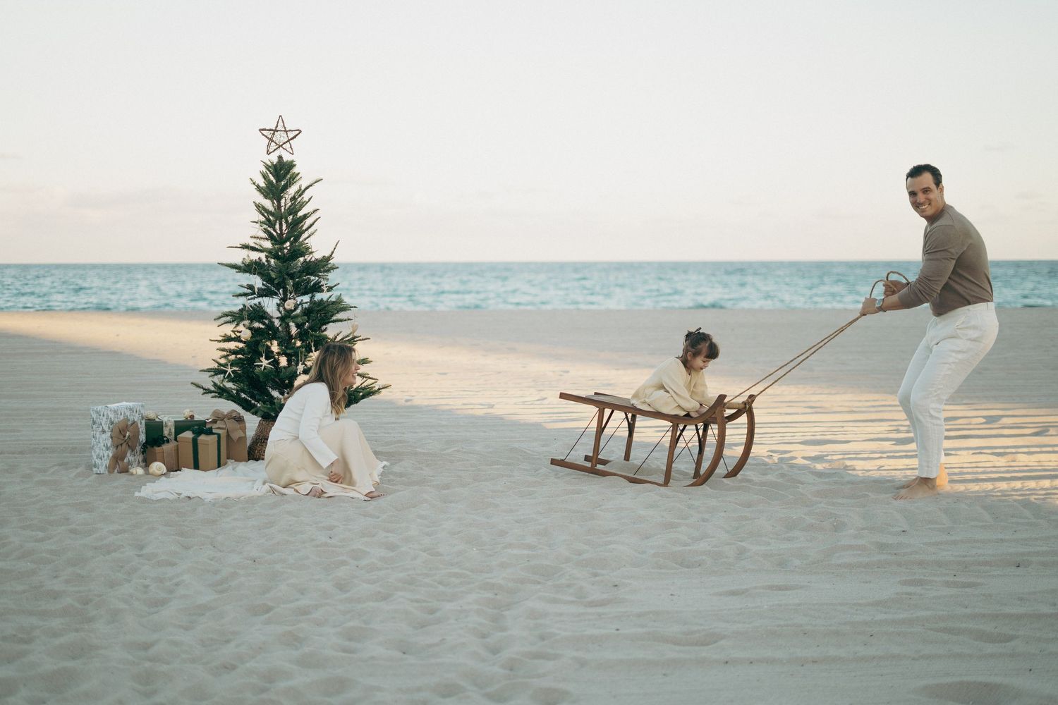Family celebrating Christmas on a sandy beach with a decorated tree, gifts, and a wooden sled at sunset.