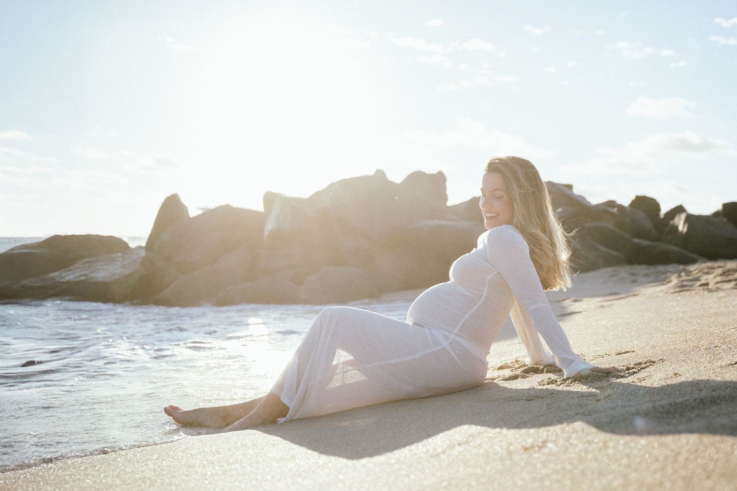 A couple in white clothing embraces on a sandy beach during golden hour as sunlight creates a warm, romantic glow.