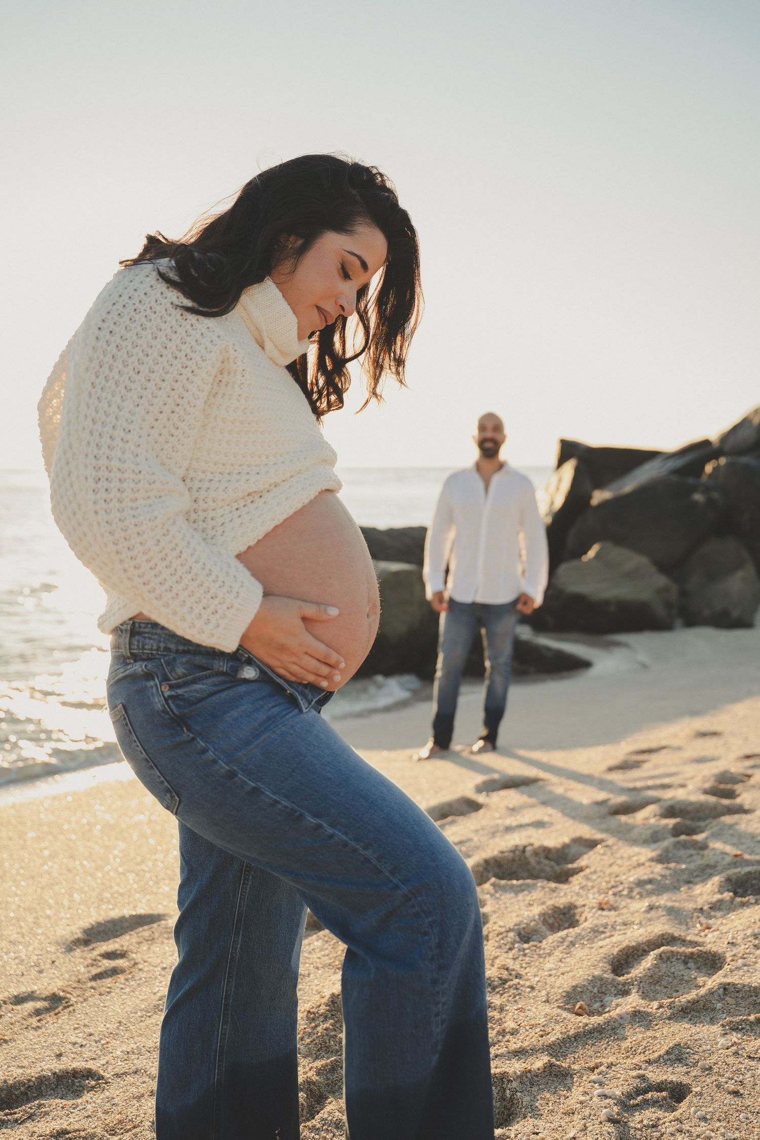 Pregnant woman in white sweater and jeans holds her belly on a sunny beach, with a man standing in the background.