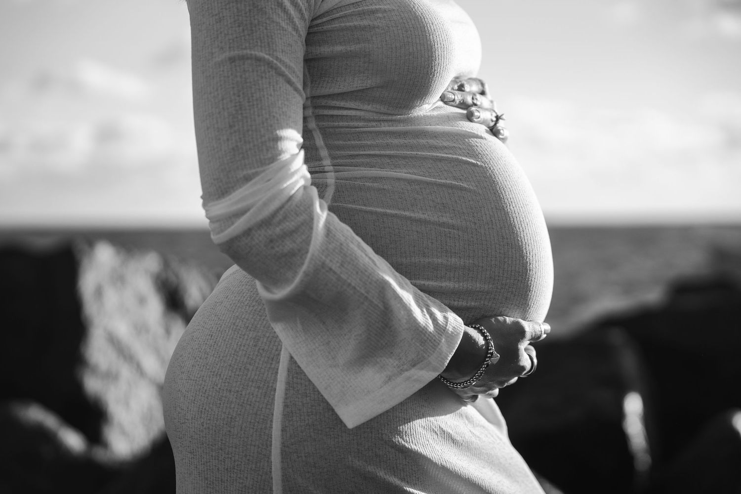 Black and white close-up photo of a pregnant belly with hands gently cradling the bump against a blurred outdoor background.