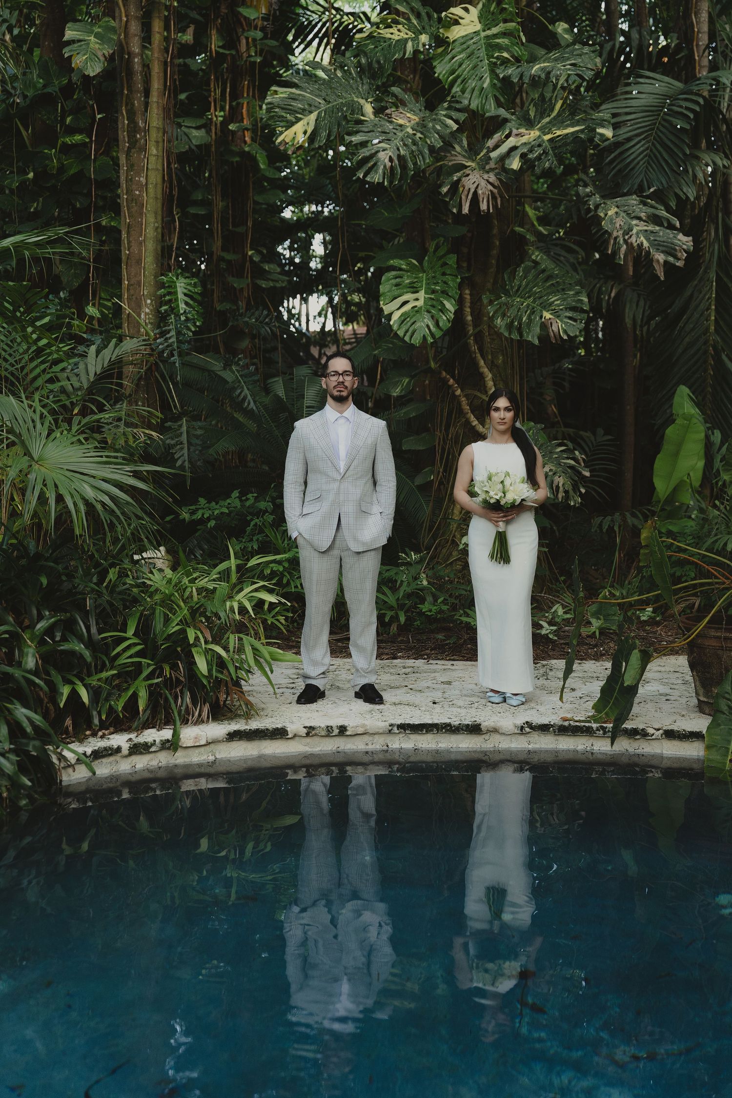 Bride and groom stand by a reflective pool surrounded by lush tropical greenery in an elegant outdoor wedding setting.