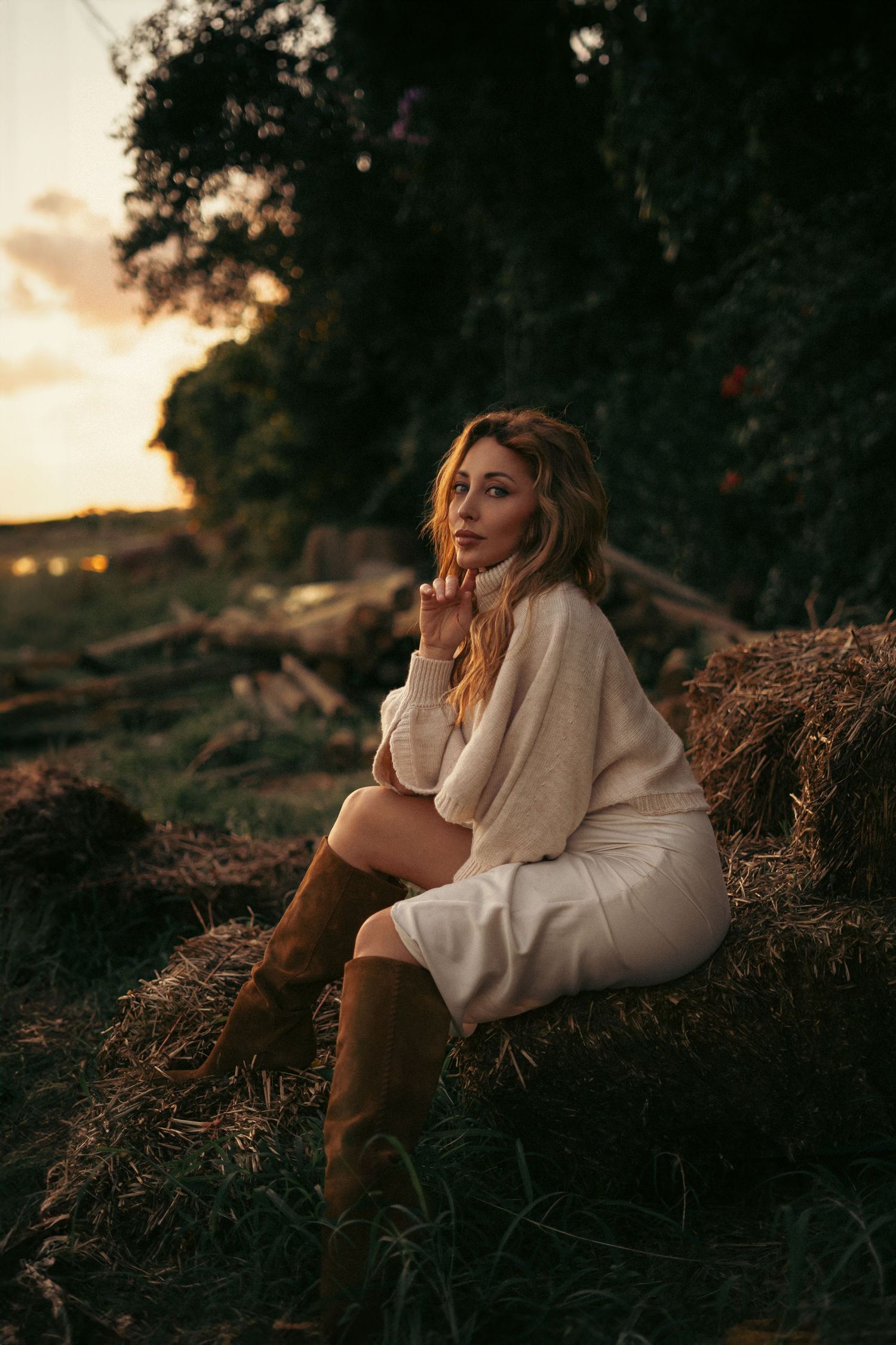 Atmospheric evening portrait on hay bales wearing white sweater and boots.