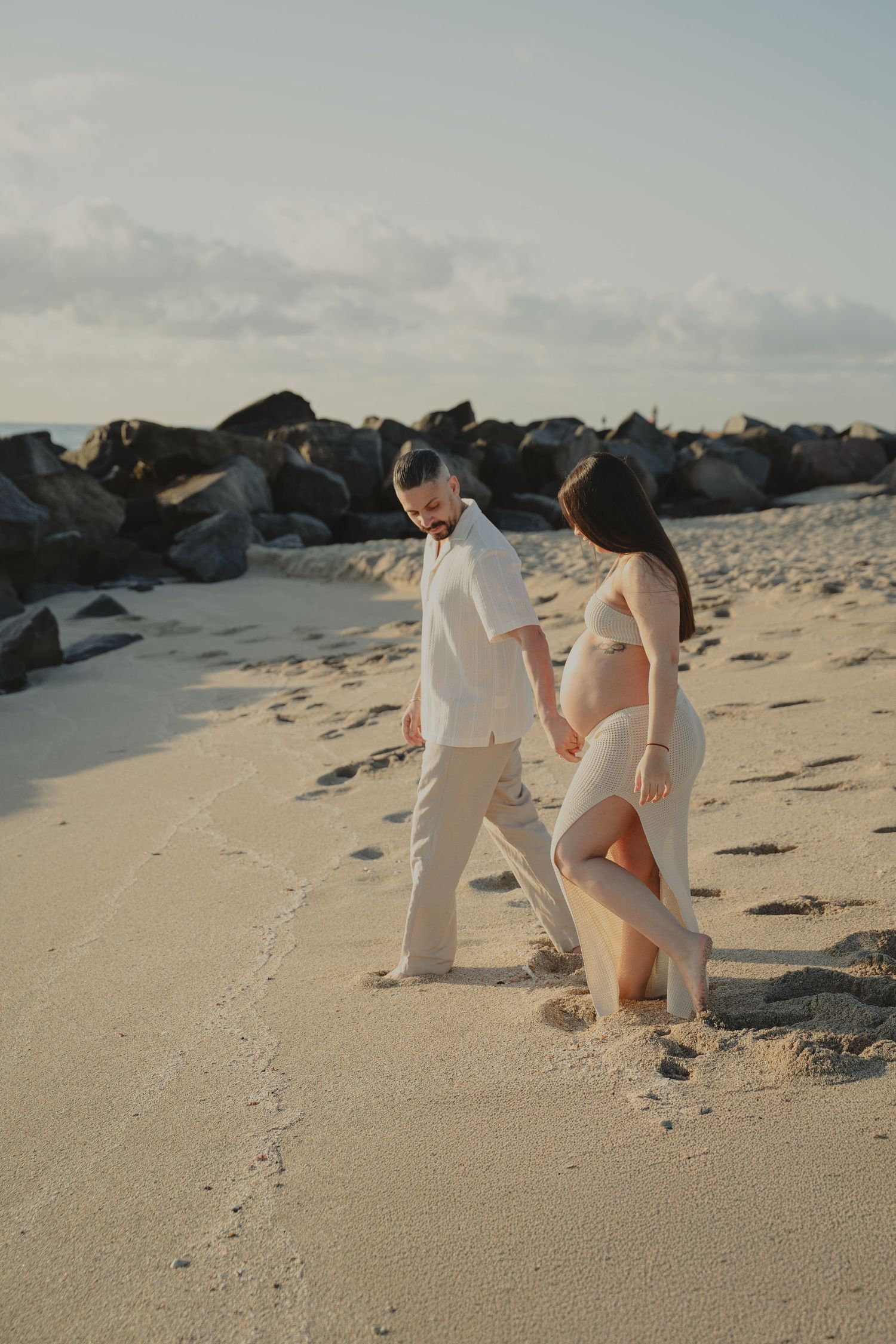 Couple in white outfits walking and playing on sandy beach near rocky shoreline.