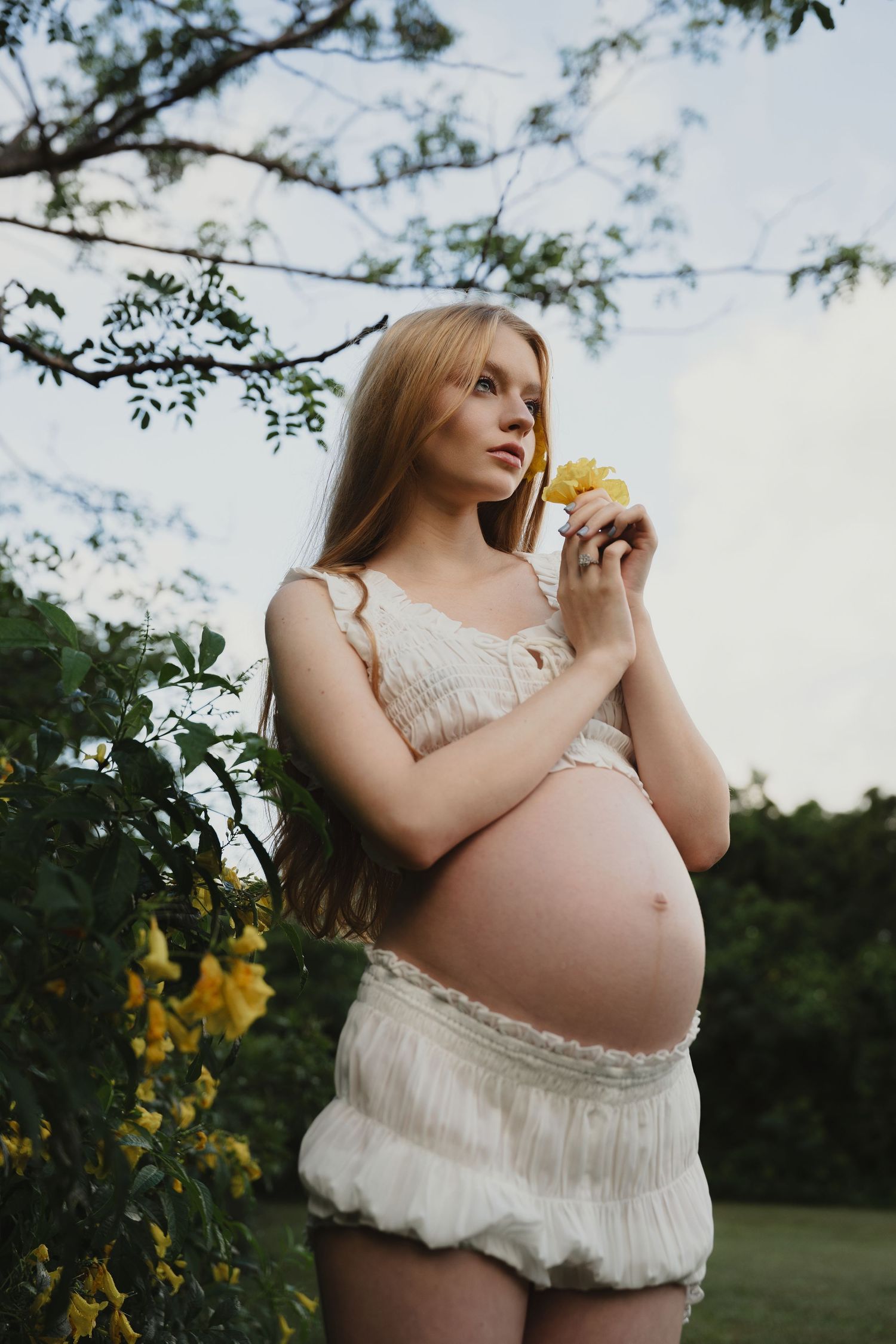 Ethereal maternity portrait in a white lace dress against a natural outdoor setting.