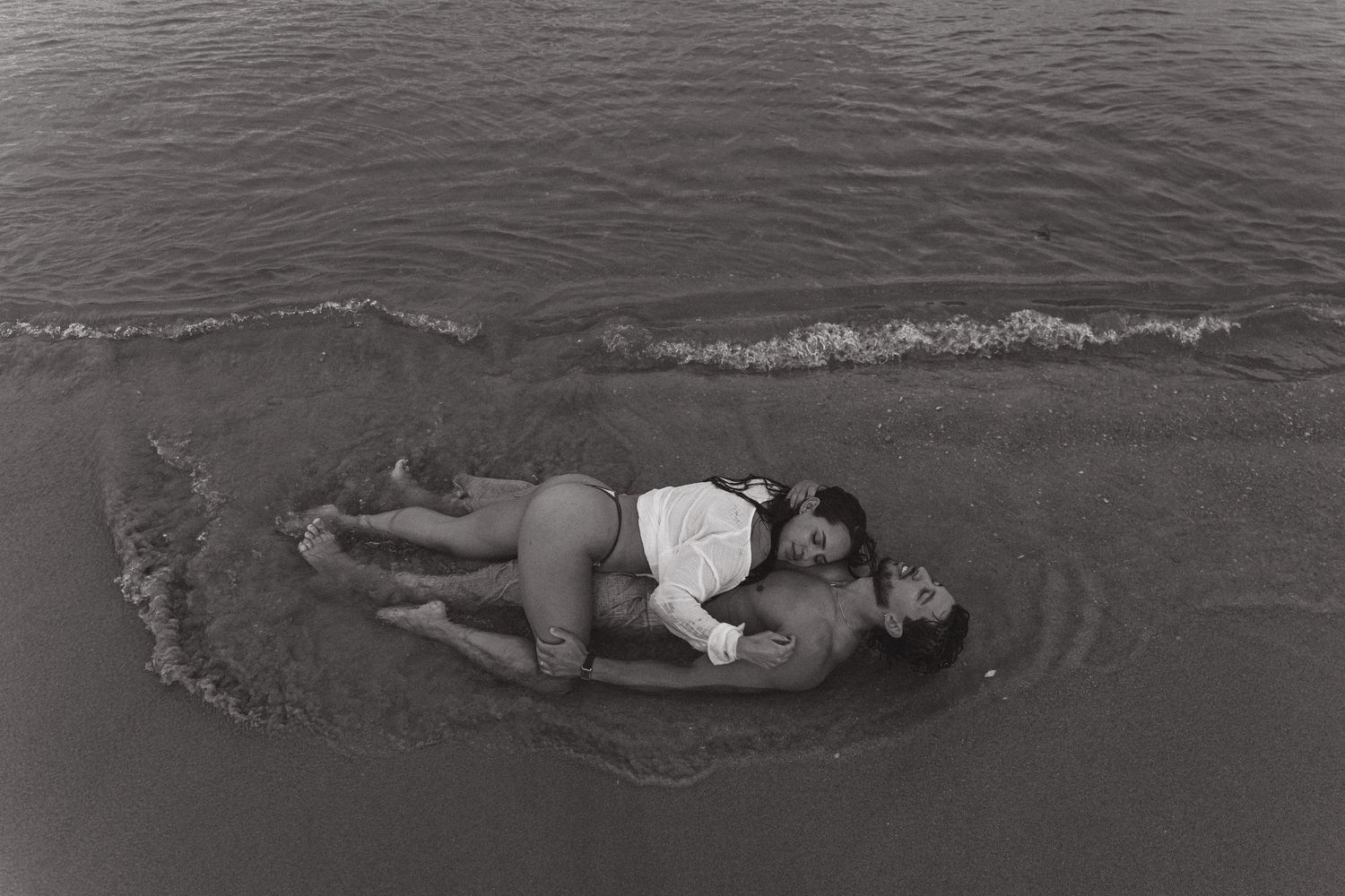 Black and white photo of two people lying together in shallow ocean waves on a beach.